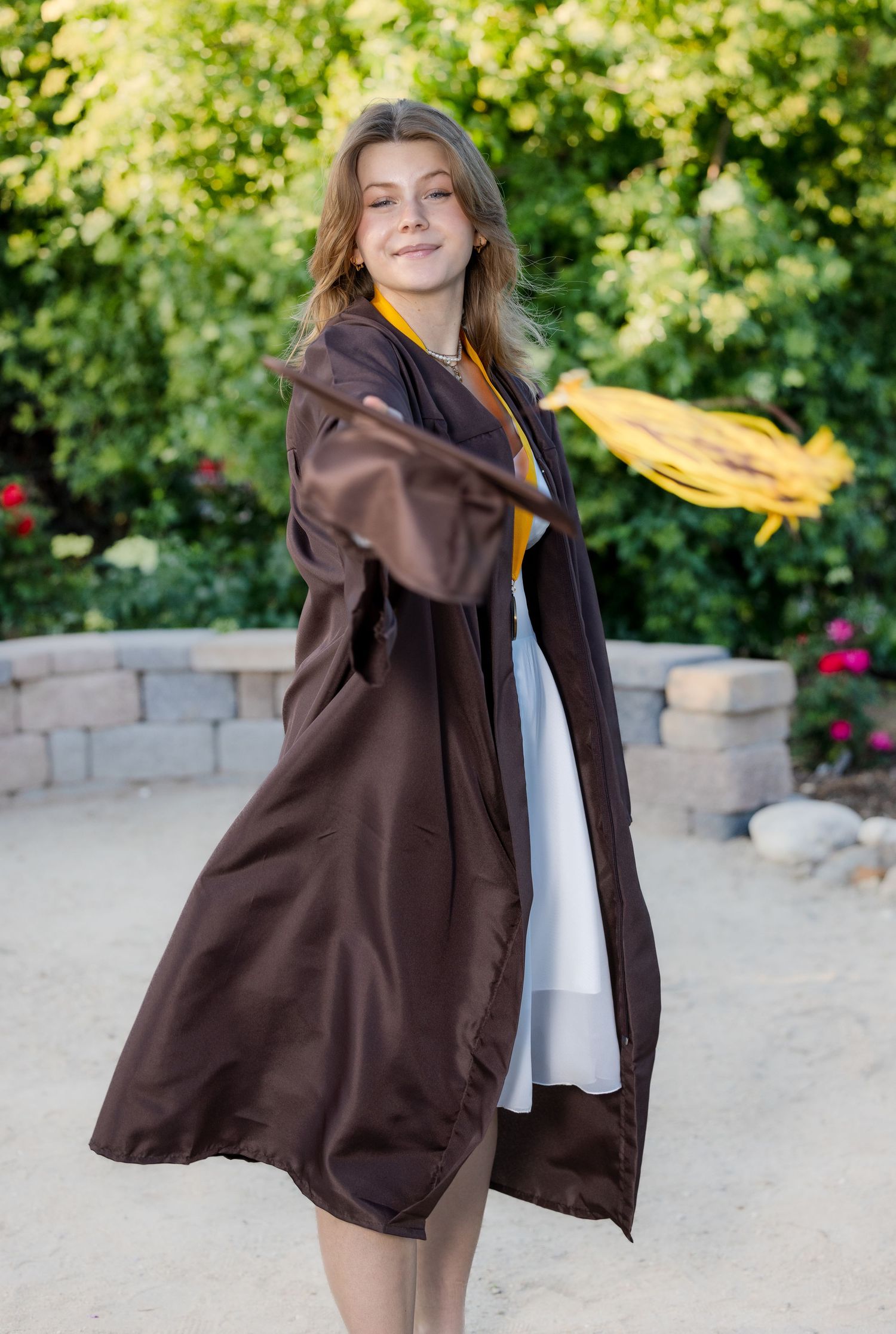 Person in brown oversized jacket and white shirt poses outdoors against stone wall and green foliage background.