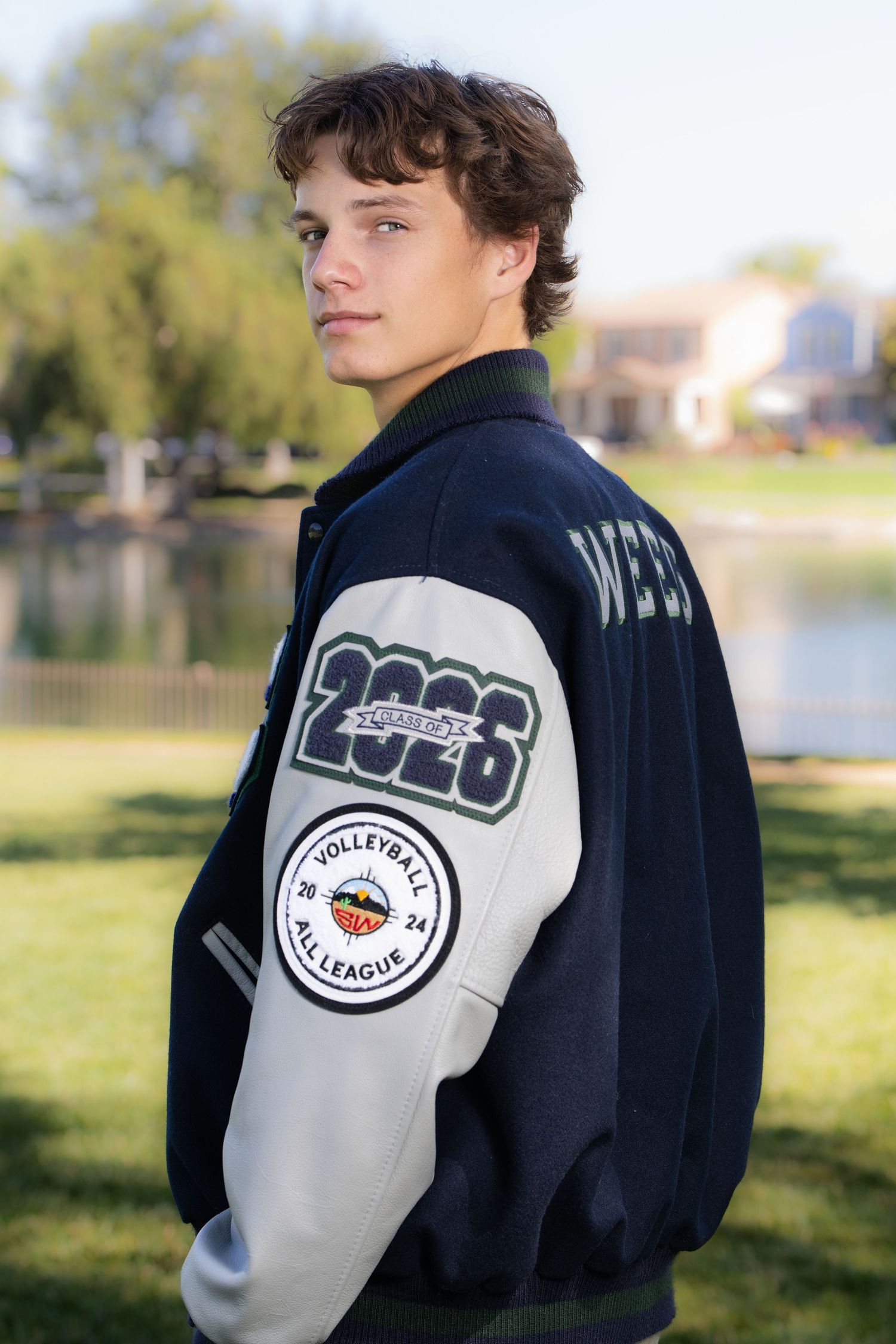 Teen boy in varsity letterman jacket with 2026 volleyball patch poses outdoors near pond.