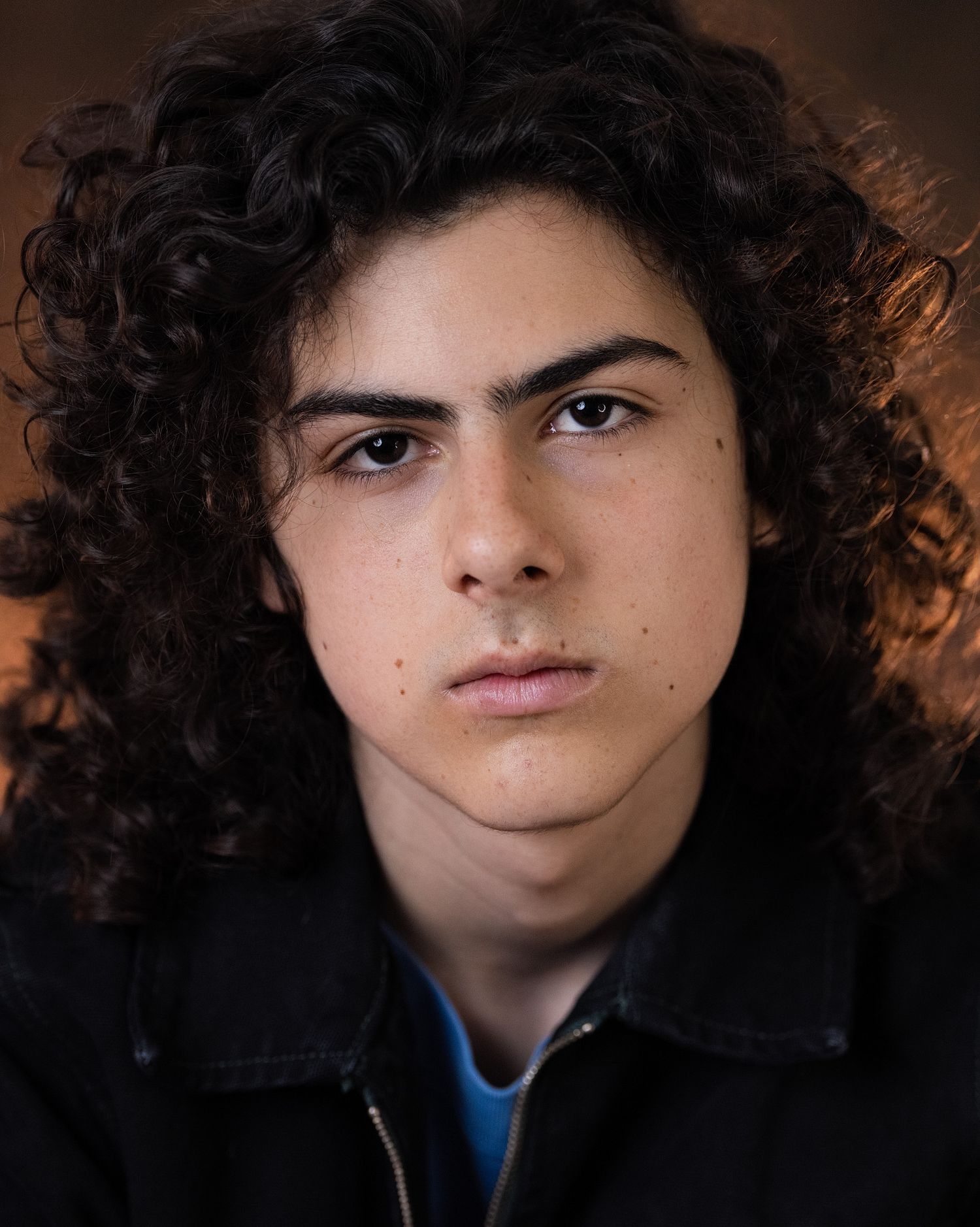 Young man with curly dark hair and bold eyebrows wearing a black jacket, captured in a dramatic close-up portrait.
