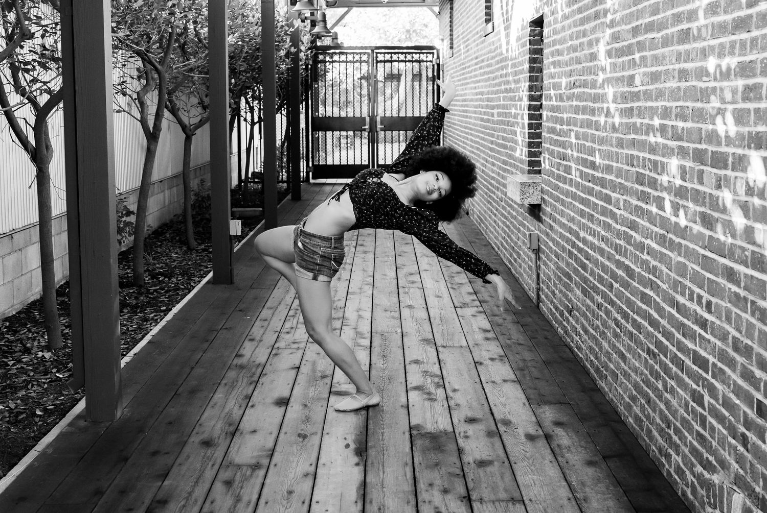 A dancer performs a graceful backbend pose on a wooden deck next to a brick wall in black and white.