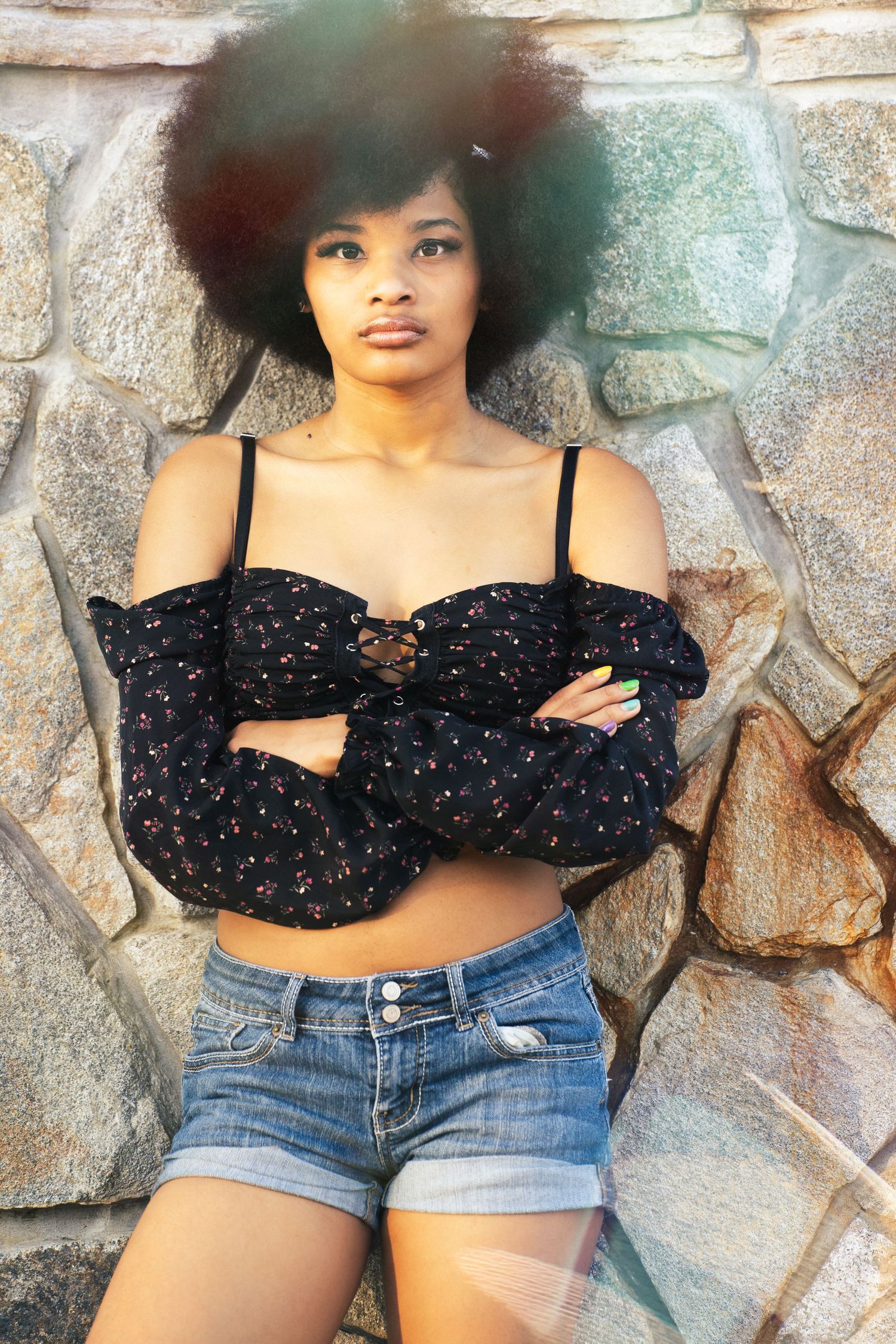 Four fashion portraits showing a stylish outfit with denim shorts and a black patterned crop top against a stone wall.