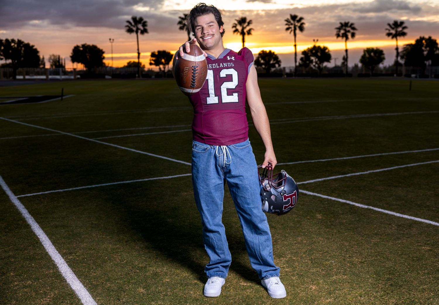 Football player stands on field at sunset with palm trees silhouetted in background.