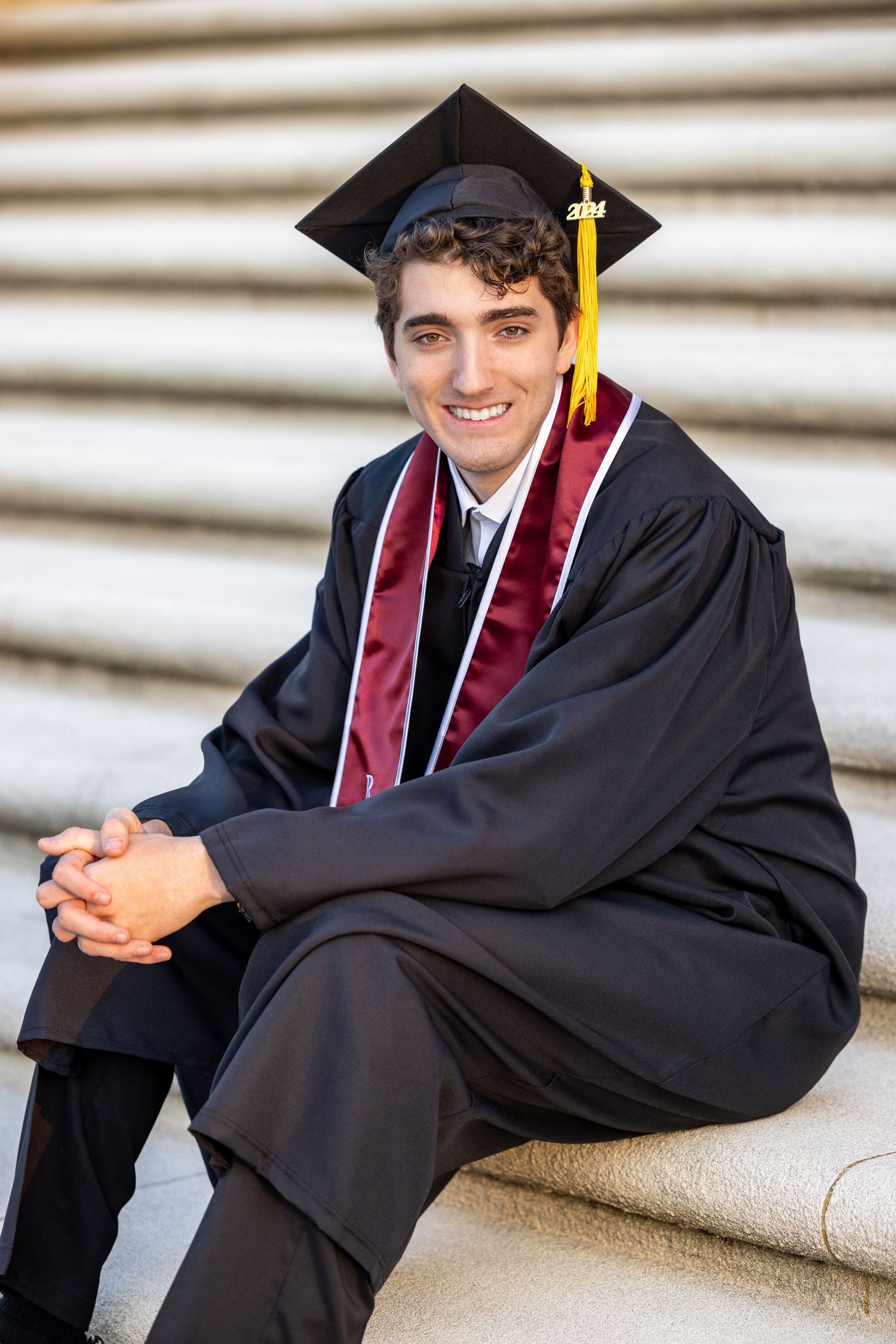 Graduate in black cap and gown with burgundy stole sits on stone steps smiling during graduation photo session.