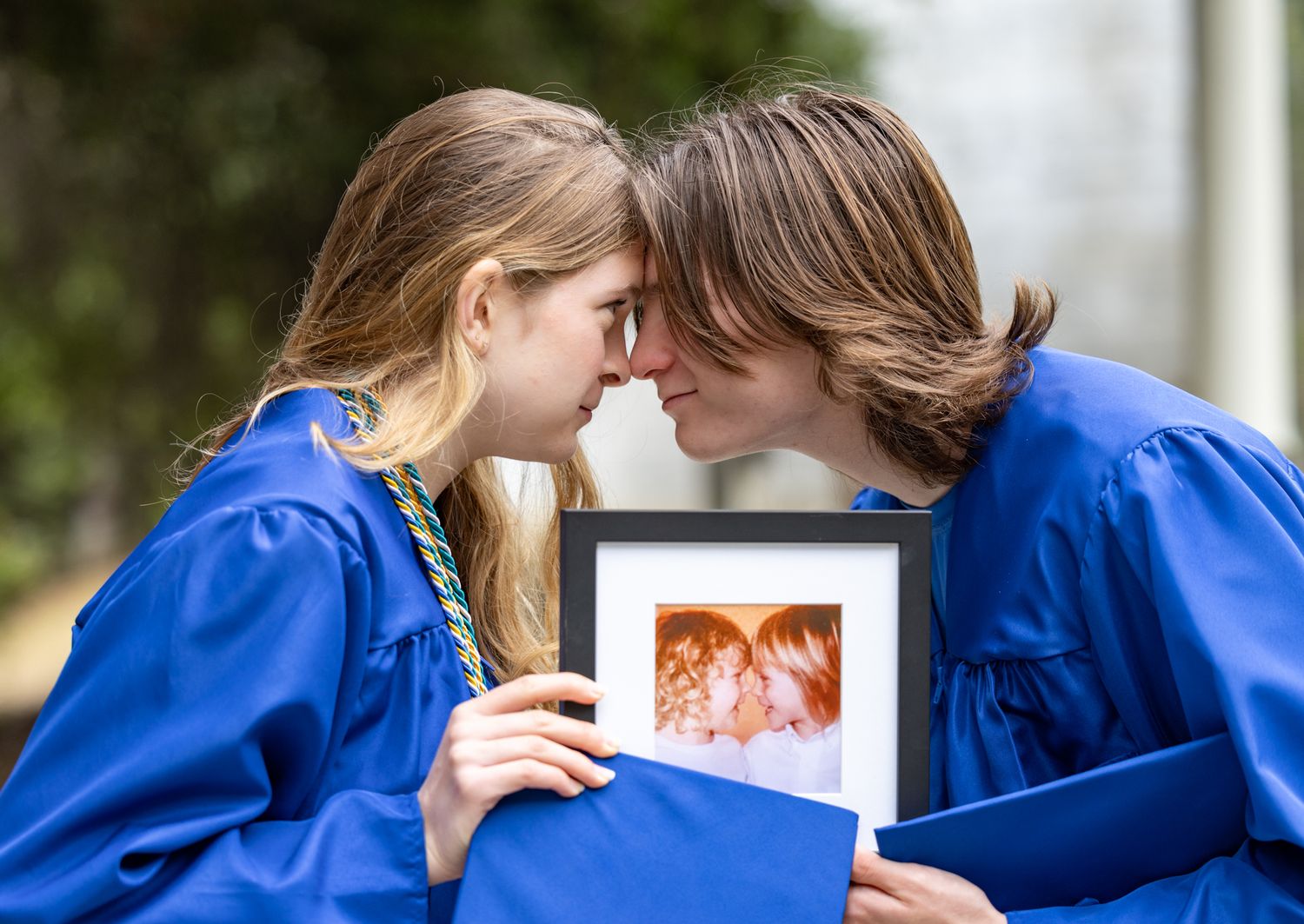 Two graduates in blue caps and gowns share a tender moment while holding a framed photograph together.