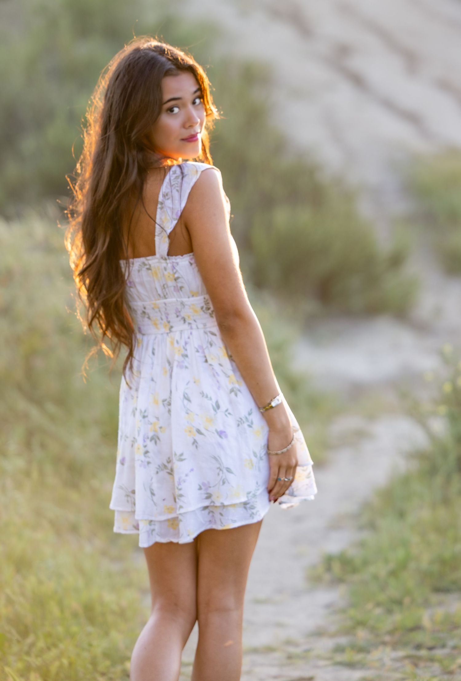 A young woman in a white floral sundress poses on a path during golden hour lighting.