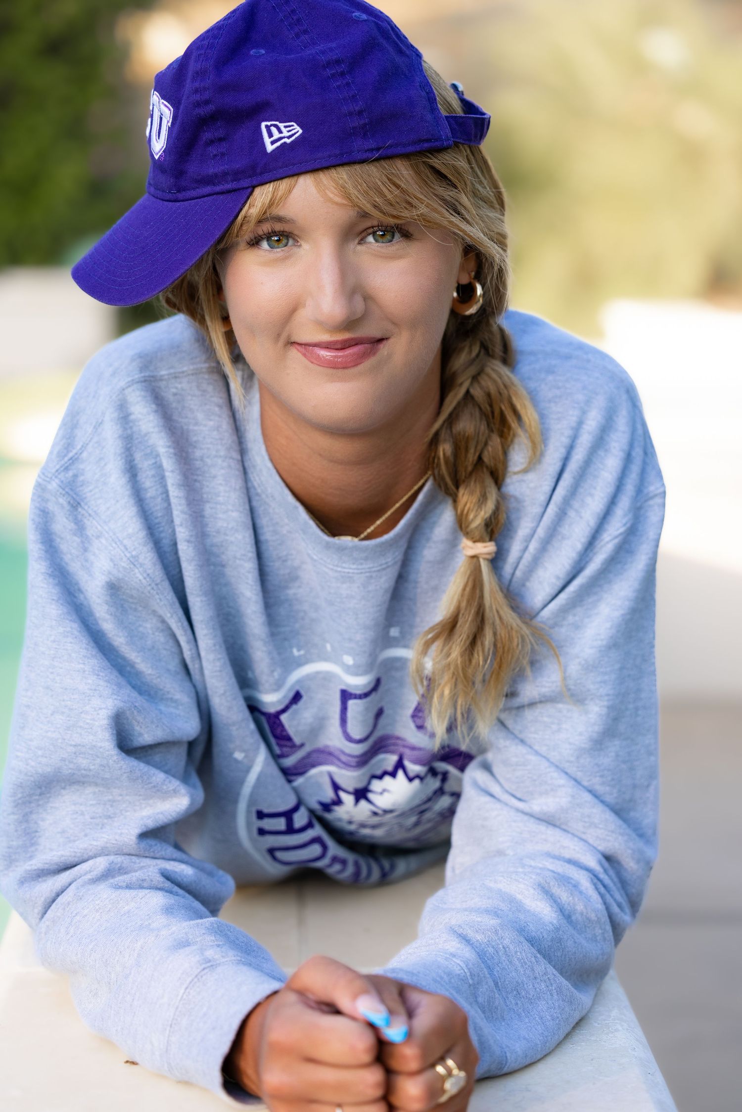 A person wearing a blue baseball cap and gray sweatshirt smiles while lying down in a casual outdoor setting.