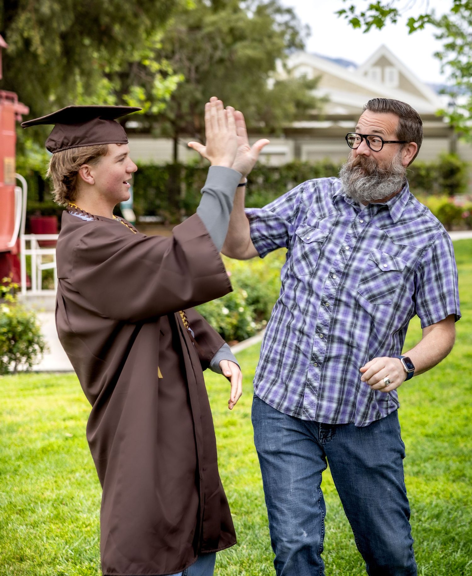 A graduate in brown cap and gown shares a celebratory high five with a bearded man in a plaid shirt on a sunny lawn.