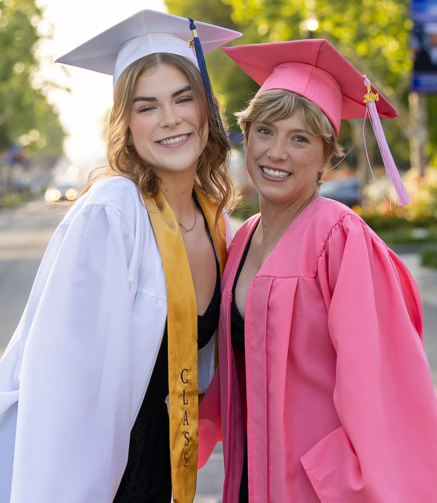 Two graduates in yellow and pink gowns share joyful moments during commencement ceremony outdoors.