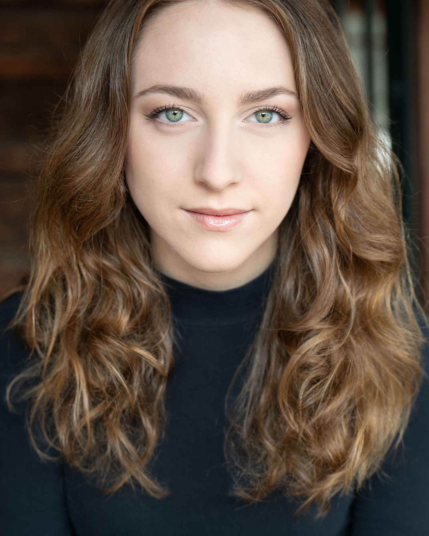 Professional headshot portrait showing soft brown wavy hair, black turtleneck, and natural makeup against dark background.