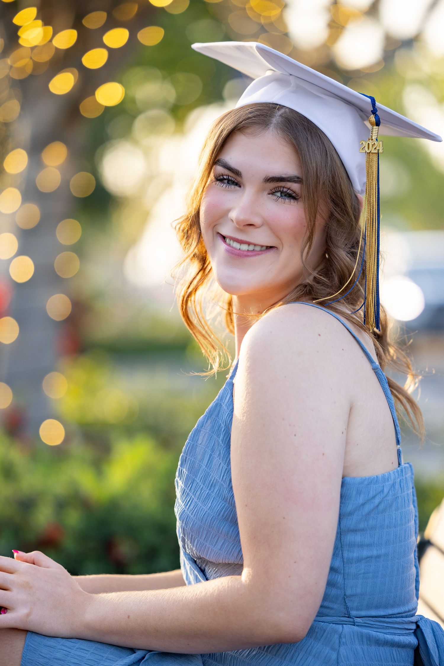 A graduate in a blue dress and cap poses outdoors with soft bokeh lights in the background during golden hour.