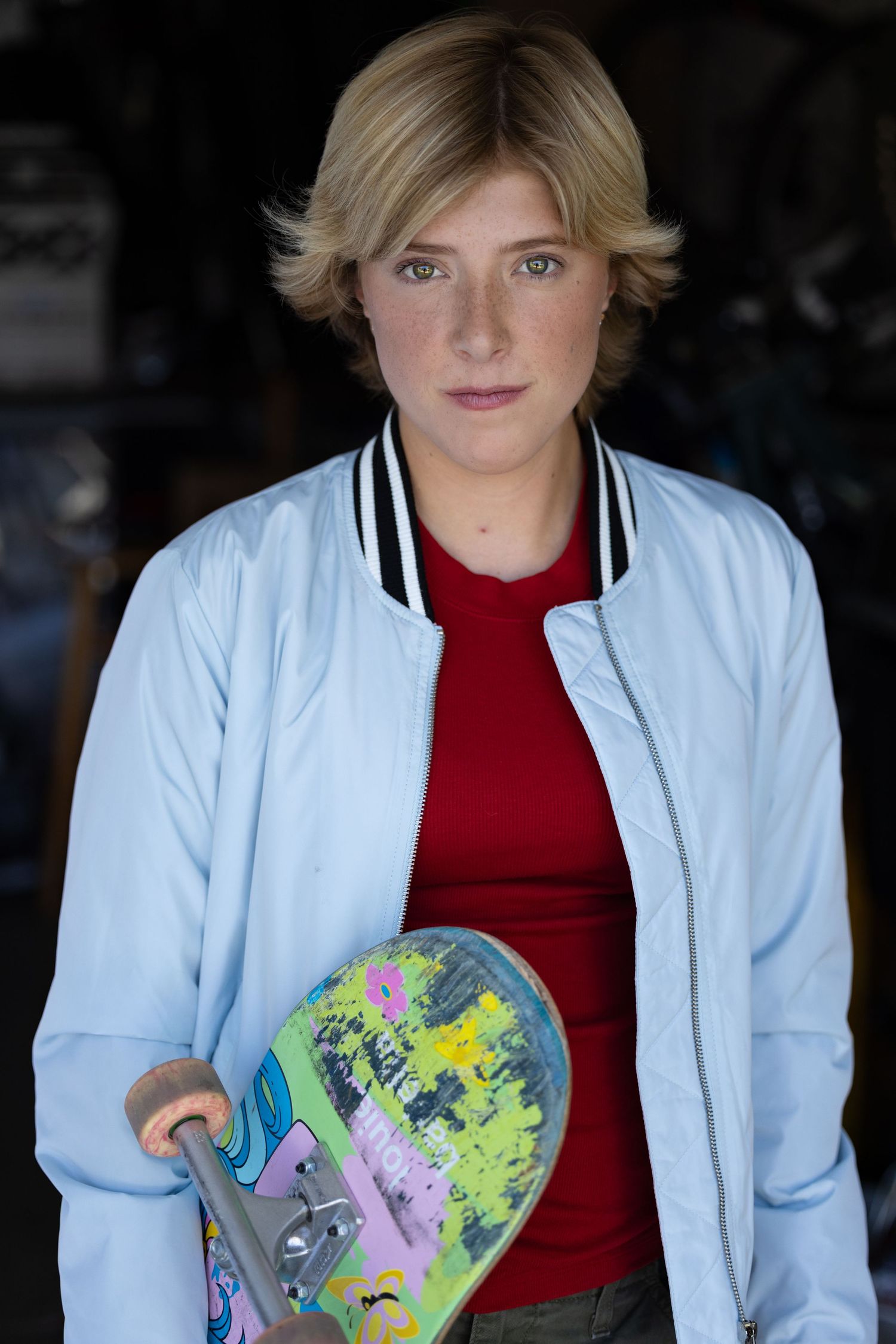 headshot for skater girl. Person in light blue jacket and red shirt holding colorful skateboard.