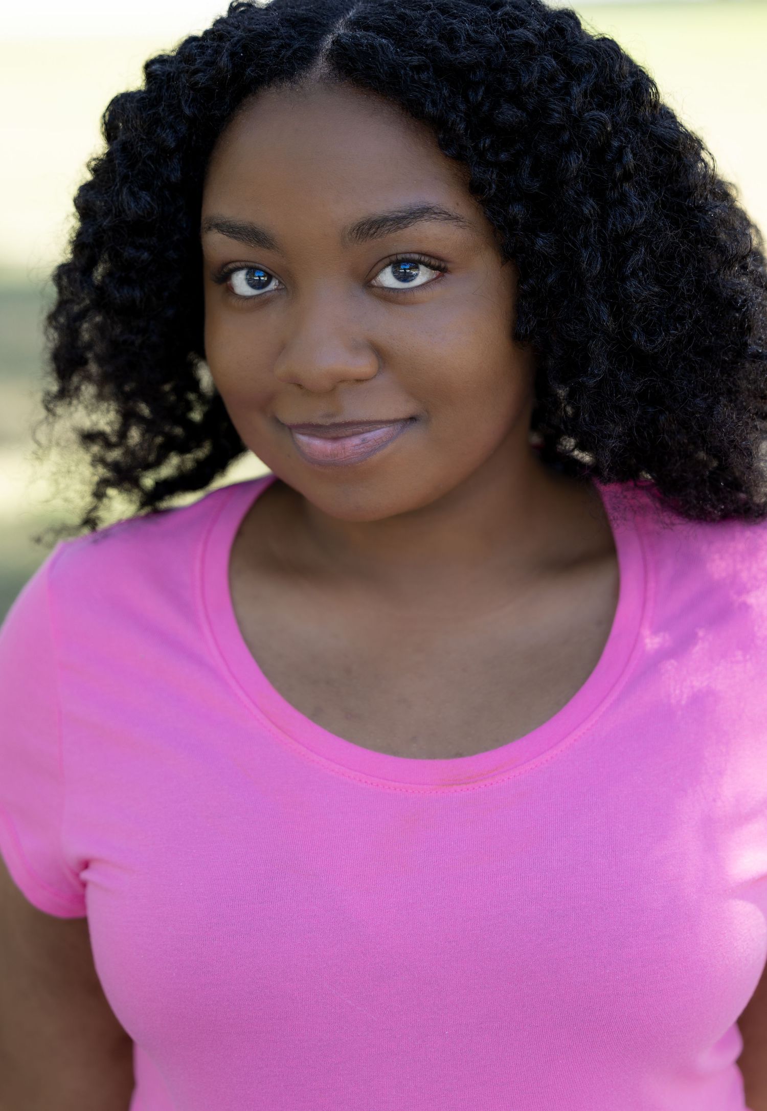 Headshots in pink top featuring bright smile and curly dark hair against neutral backdrop.