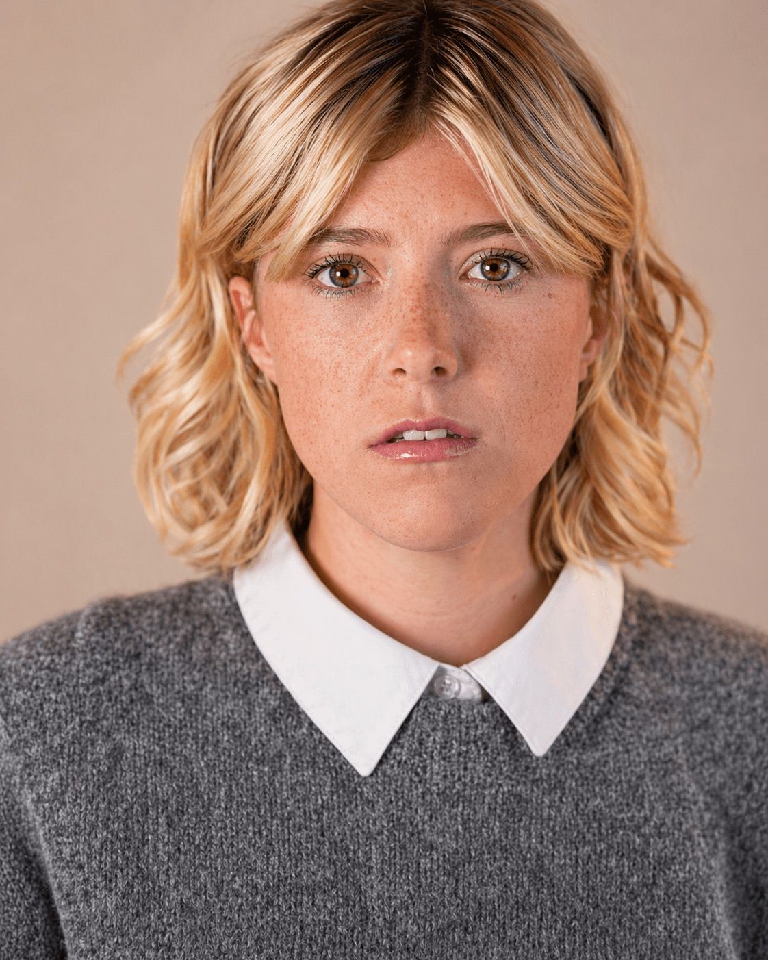 Young woman with blonde wavy hair and freckles wearing a gray sweater with white collar against neutral background.