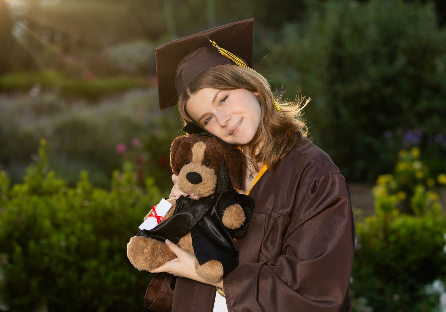 A person in a brown jacket poses playfully with a stuffed animal monkey toy in an outdoor garden setting.