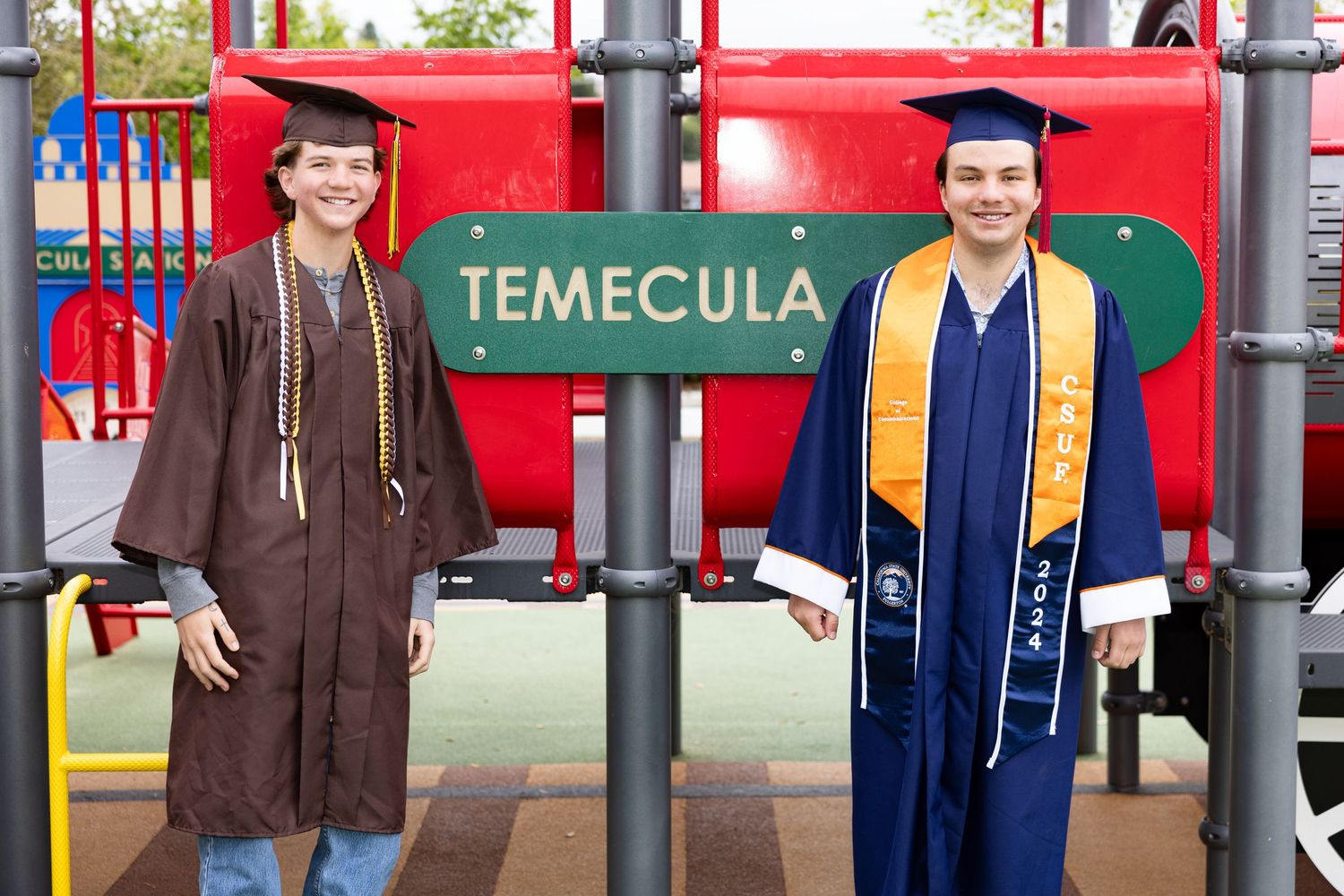 A group of graduates in blue and gold robes pose together in front of a red wall with Temecula signage.