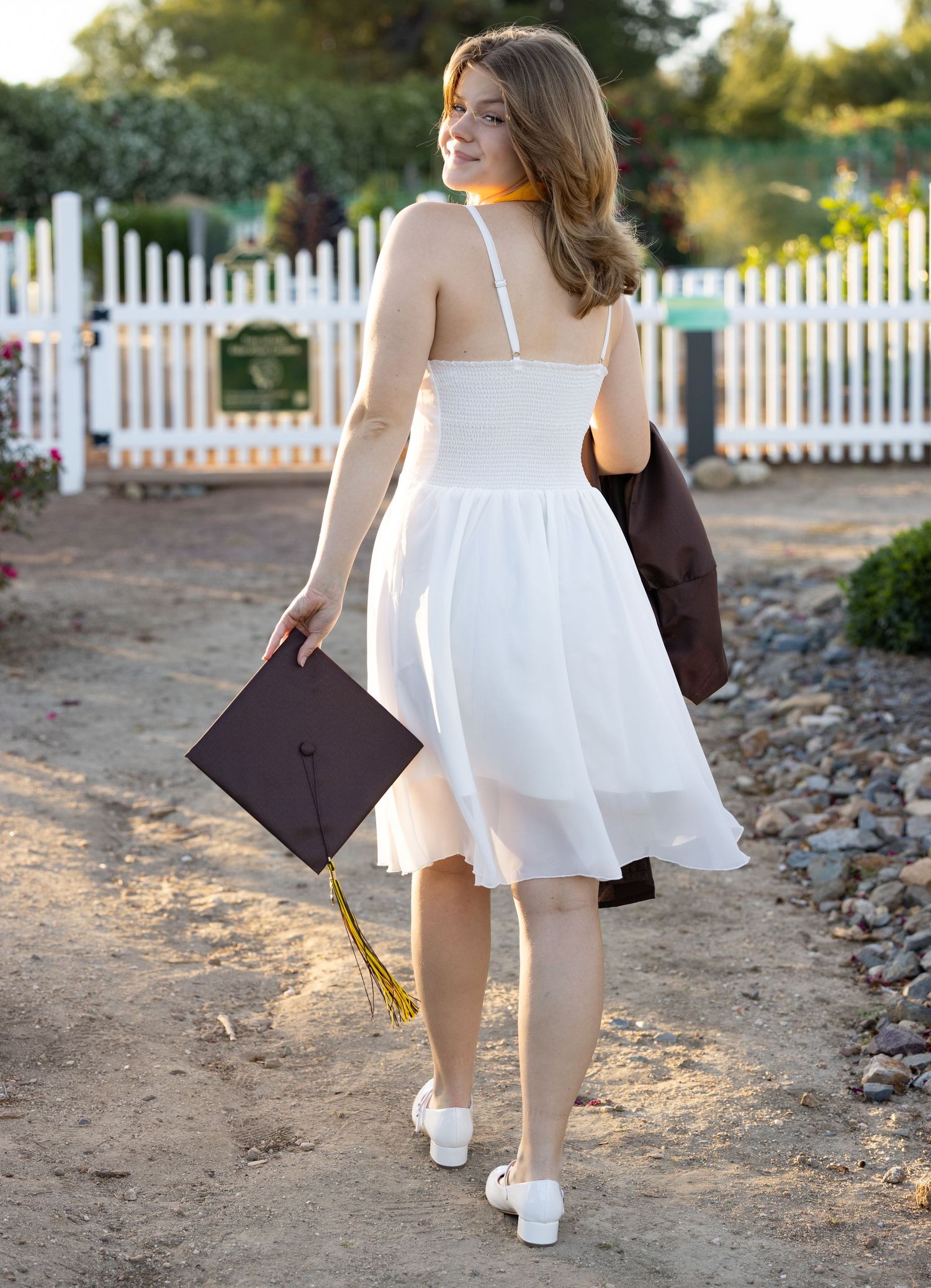 Graduate in white sundress walks away holding diploma and brown bag near white picket fence at sunset.