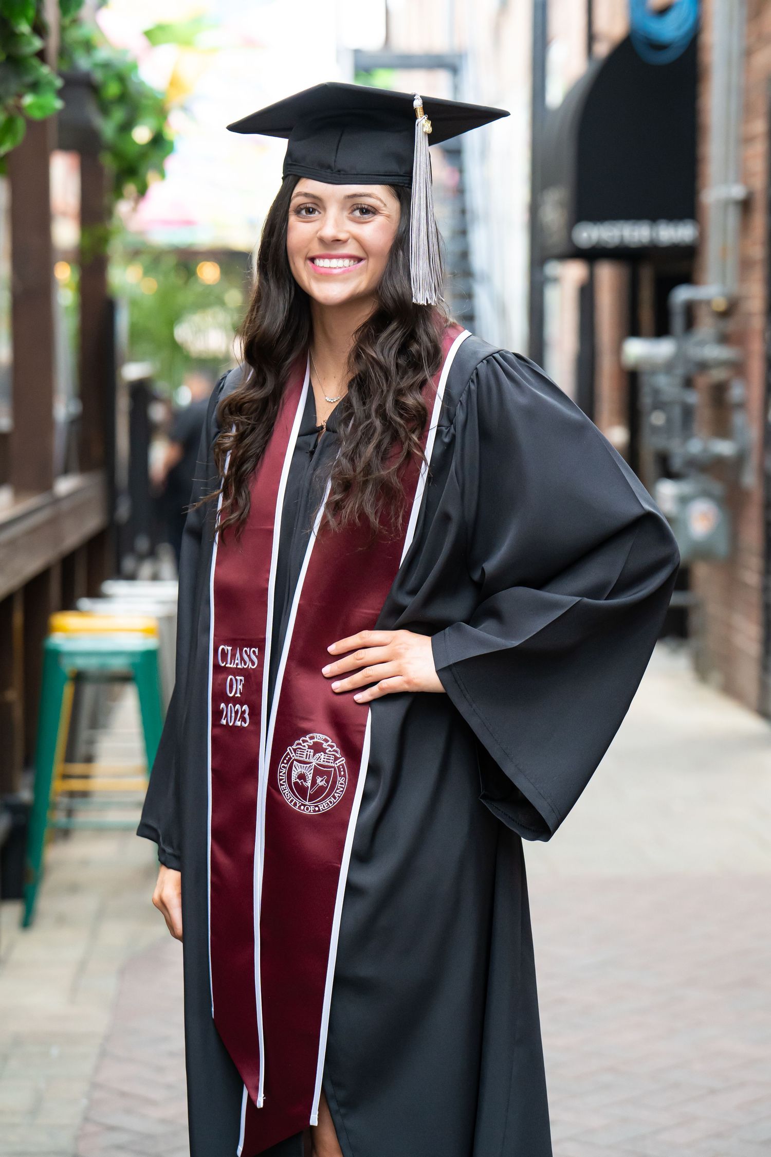 Graduate in maroon and black academic regalia poses outdoors near brick buildings during commencement celebrations.