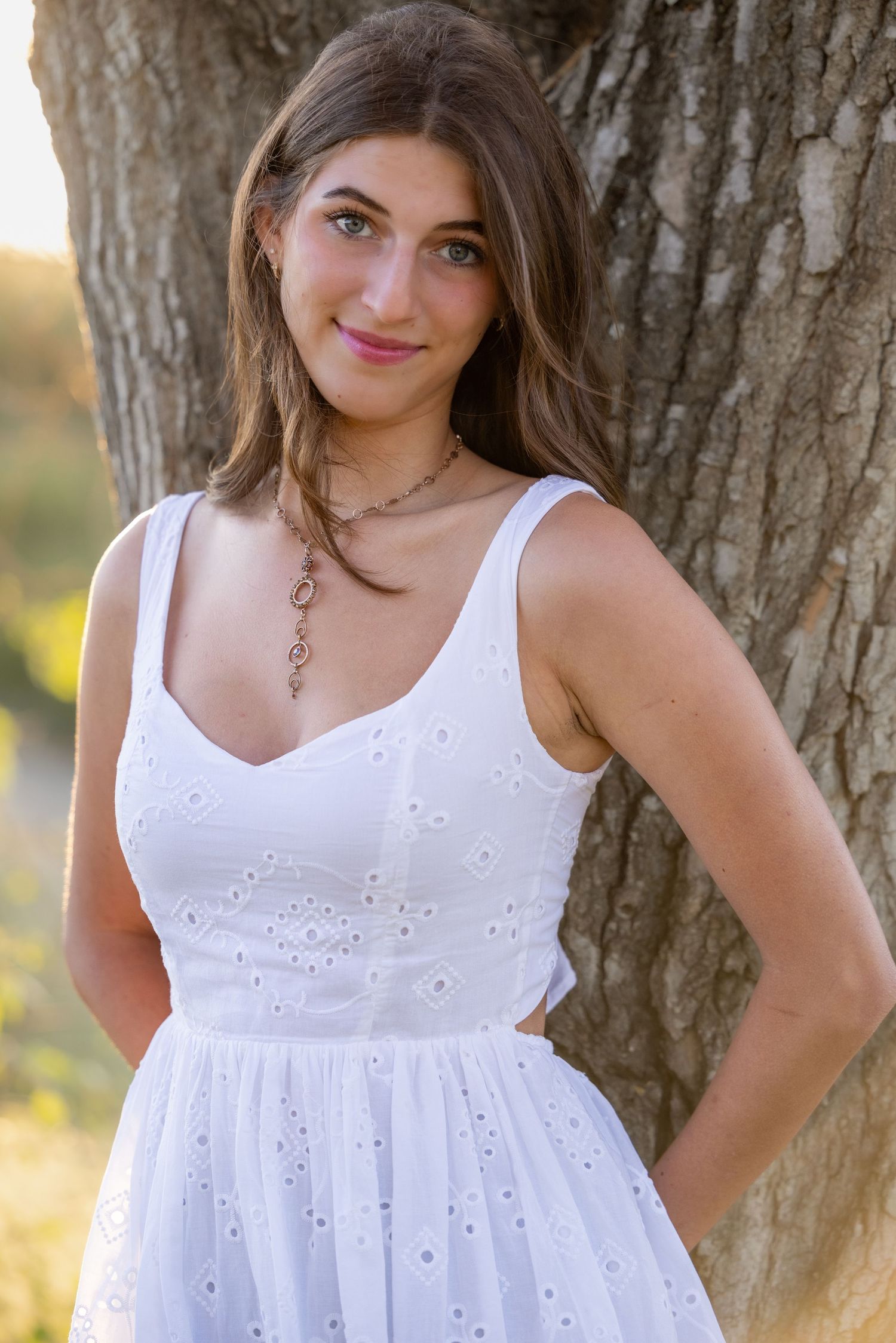 A person in a white eyelet sundress poses against a tree trunk in warm outdoor lighting.