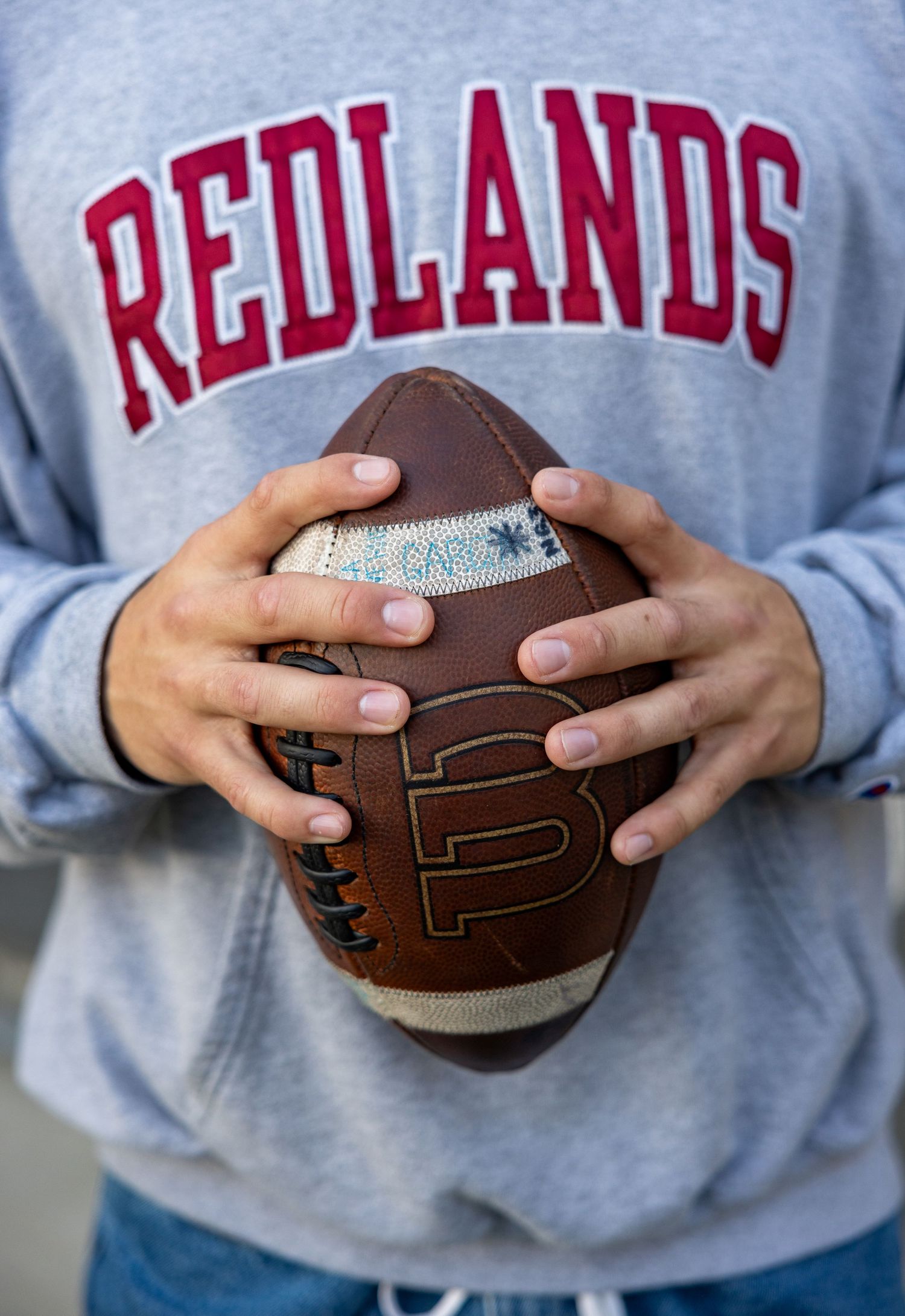 Five sequential images showing hands holding a football while wearing a grey Redlands sweatshirt.