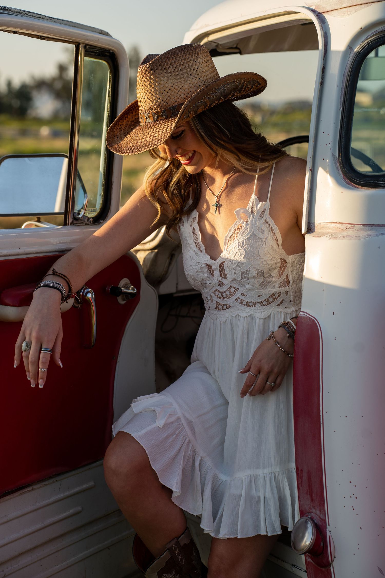 Woman in white lace dress and straw hat poses elegantly against vintage car in romantic countryside setting.