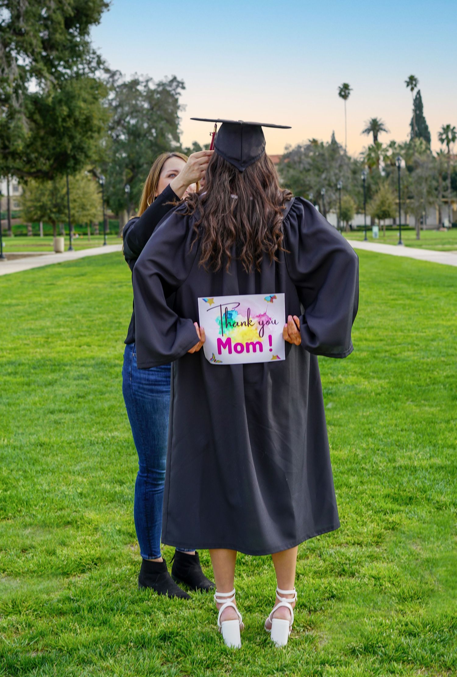 Graduate in black cap and gown stands on green lawn with sign for mom during outdoor graduation photoshoot.