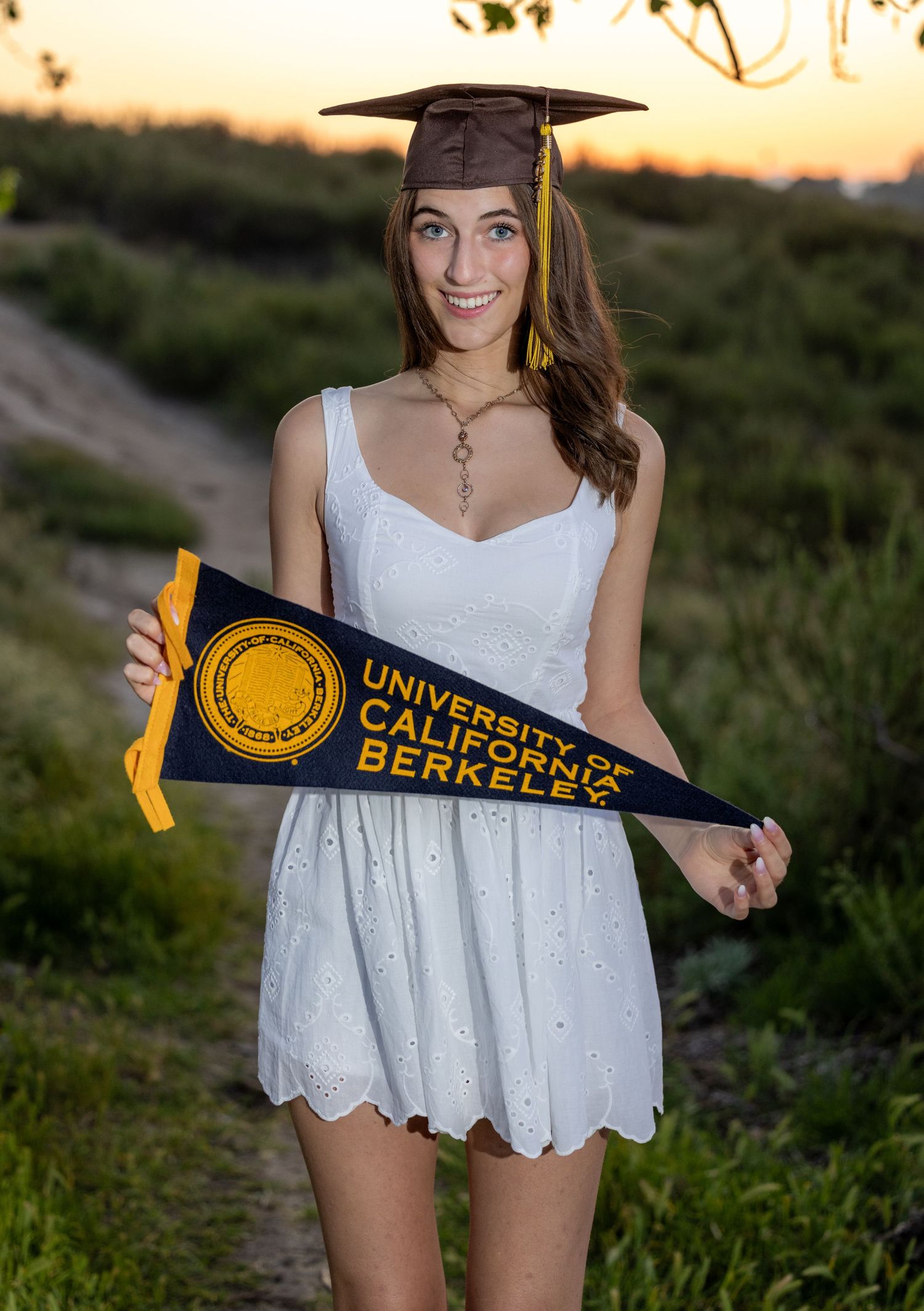 A graduate in a white dress holds a UC Berkeley pennant during sunset graduation photos outdoors.