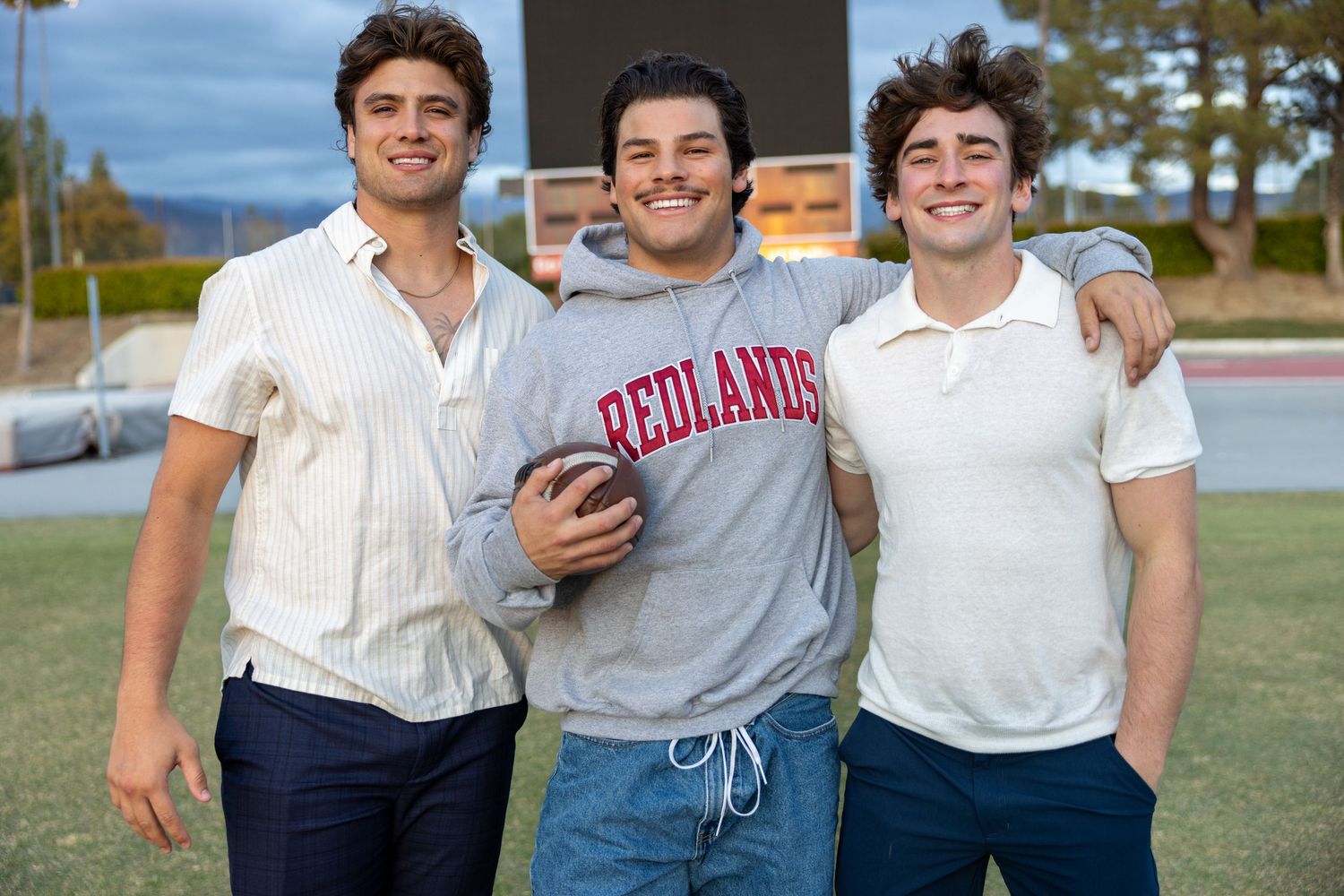 Three young men in casual attire smiling together outdoors, with one wearing a Redlands sweatshirt.
