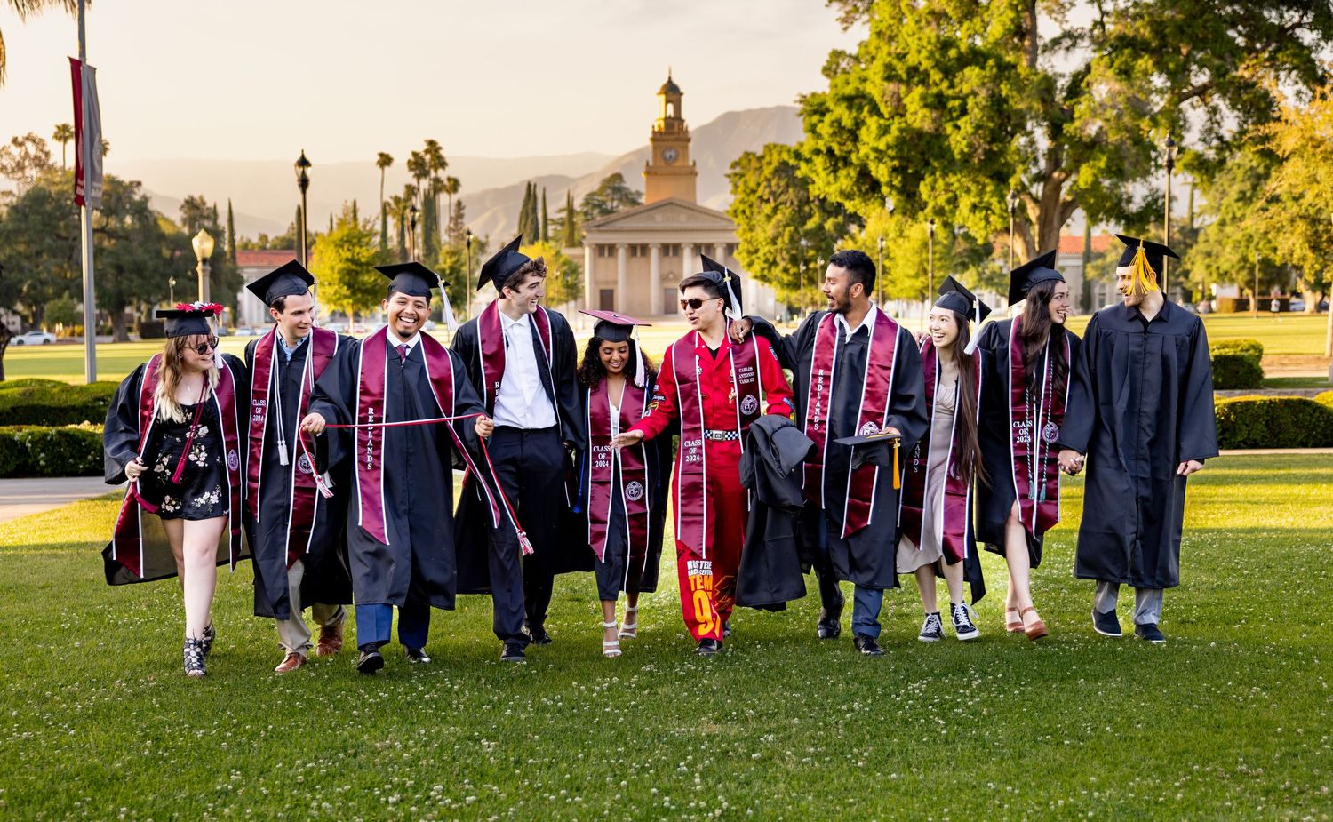Graduates in colorful academic regalia walk together on a sunny campus lawn with trees and buildings in the background.
