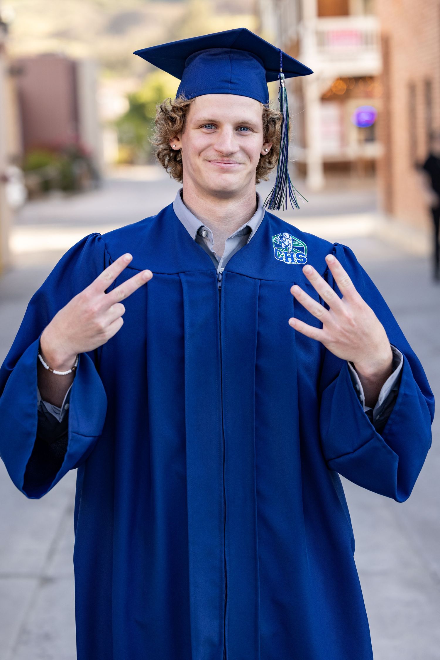 Graduate in blue cap and gown makes playful hand gestures in outdoor portrait series.