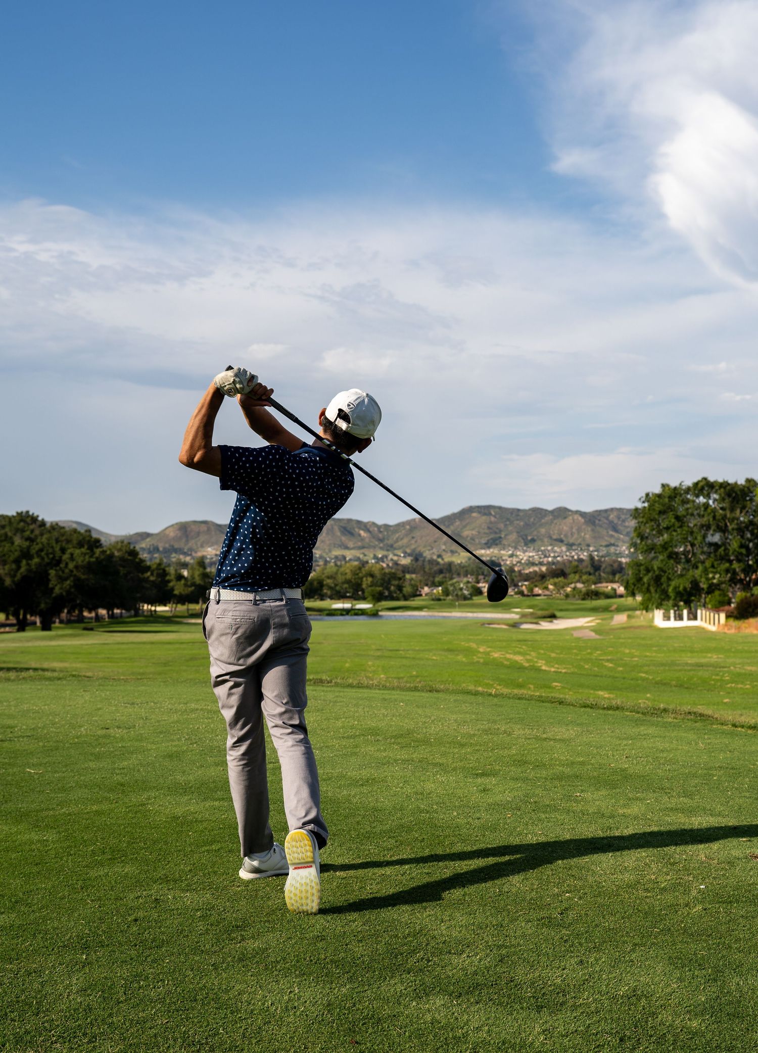 Golfer in blue polo shirt takes a swing on lush green course with mountains in background under blue sky.