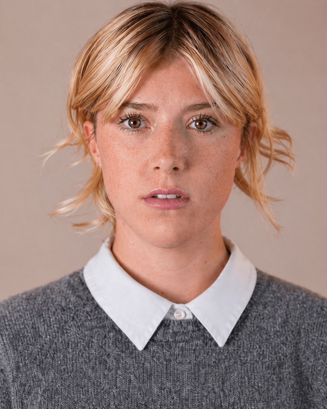 Young blonde woman with freckles wearing a gray sweater and white collared shirt, neutral studio headshot.