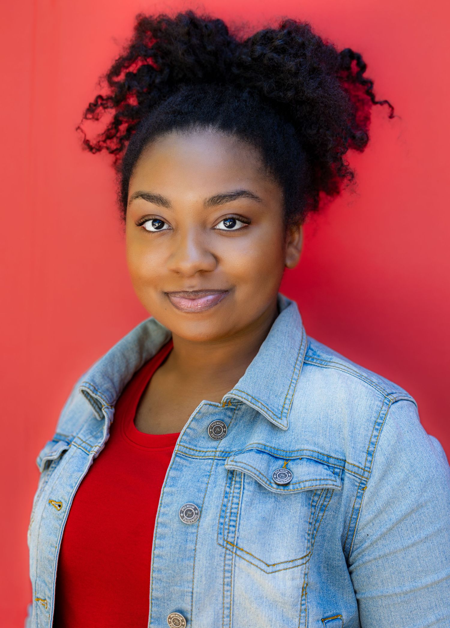 Vibrant portraits against red wall wearing denim jacket and red top with natural curls.