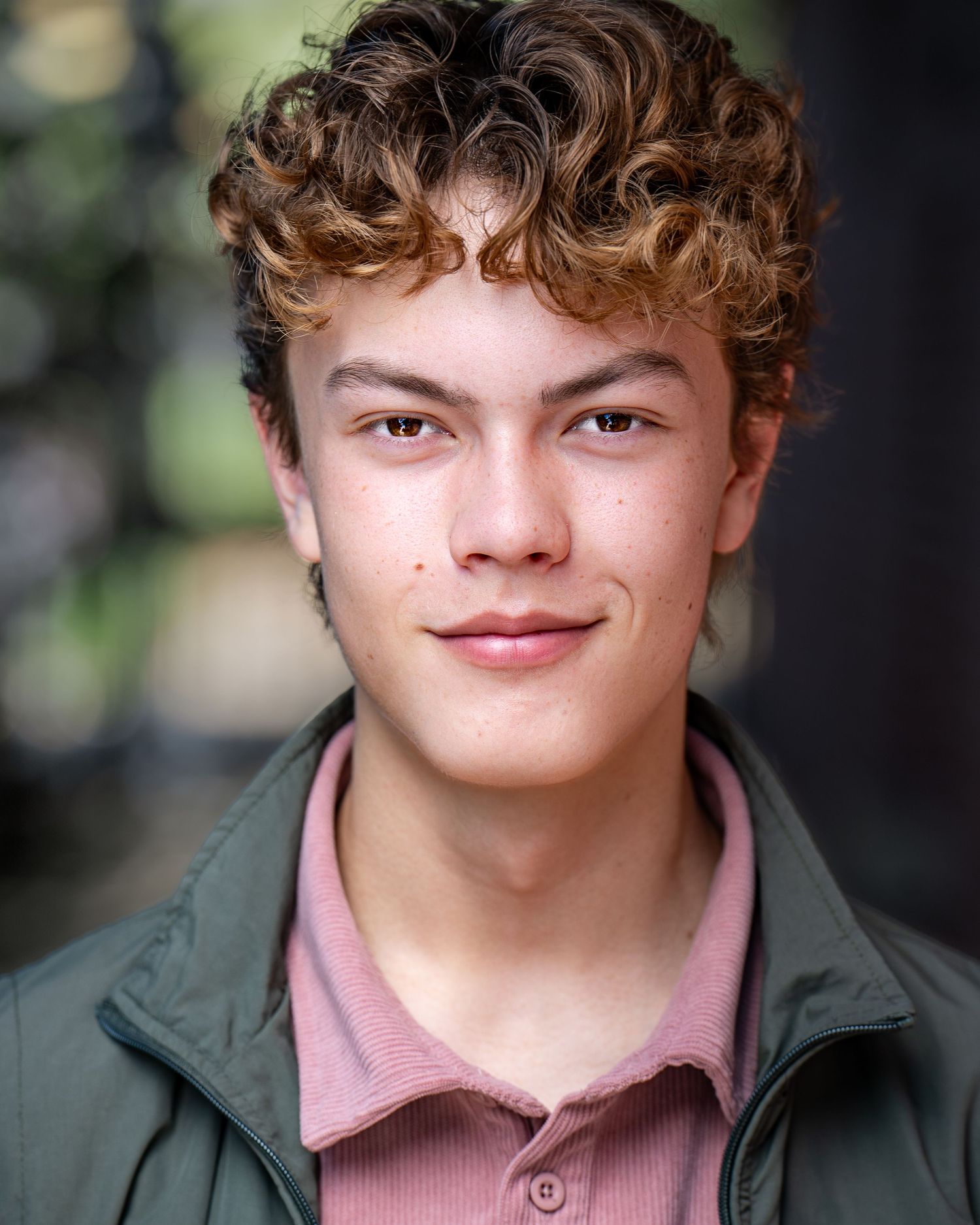 A young person headshot in a green coat and pink collared shirt smiles while outdoors in natural lighting.