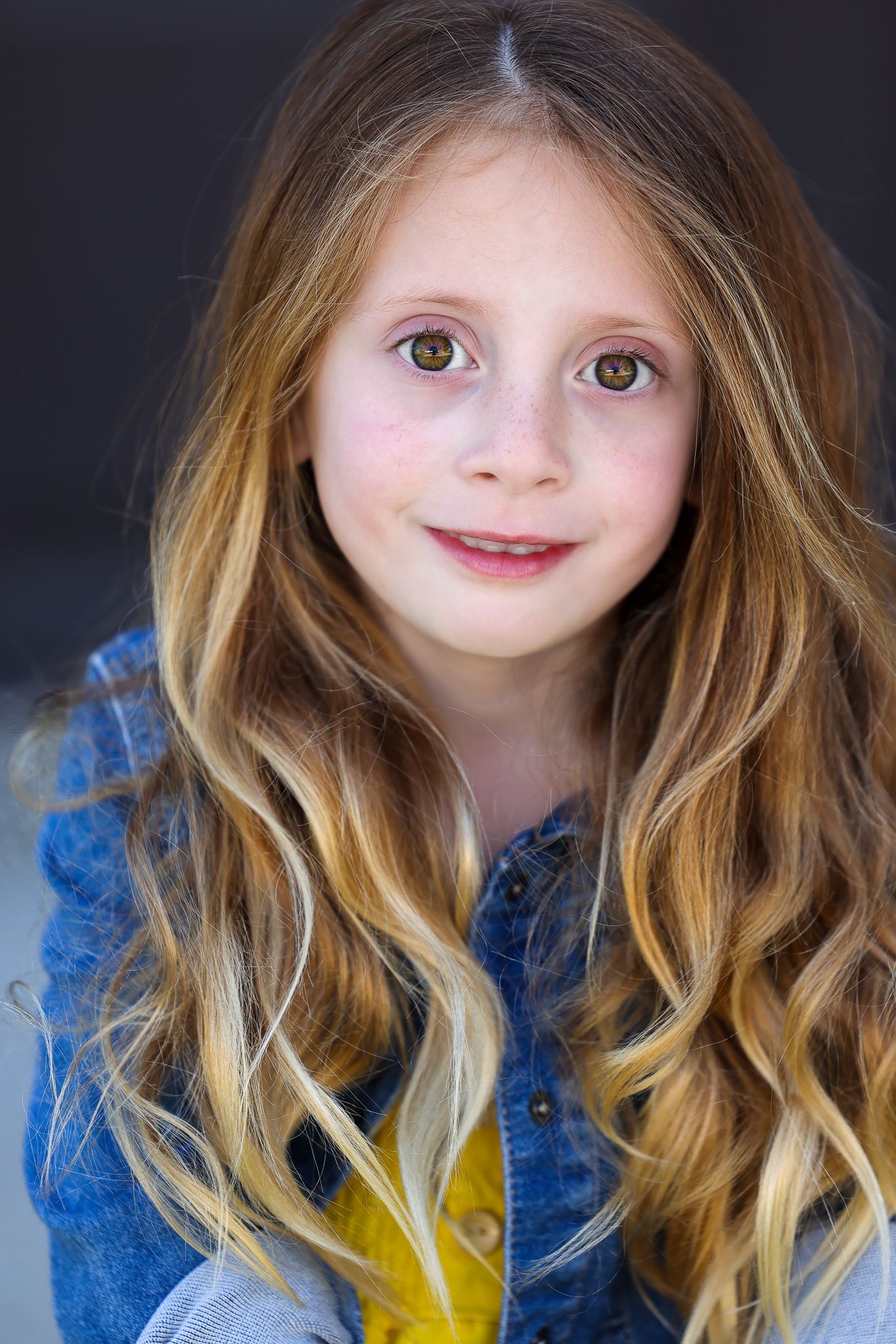Child in denim jacket and yellow shirt against dark background.