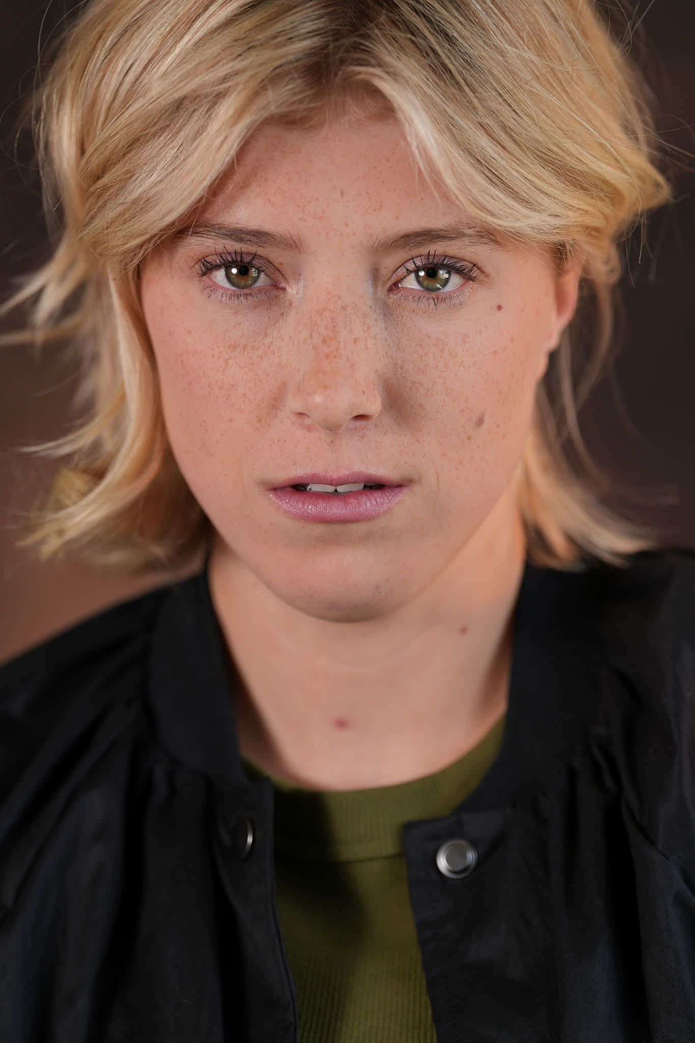 Young blonde woman with freckles and green eyes wearing a black leather jacket and olive shirt.
