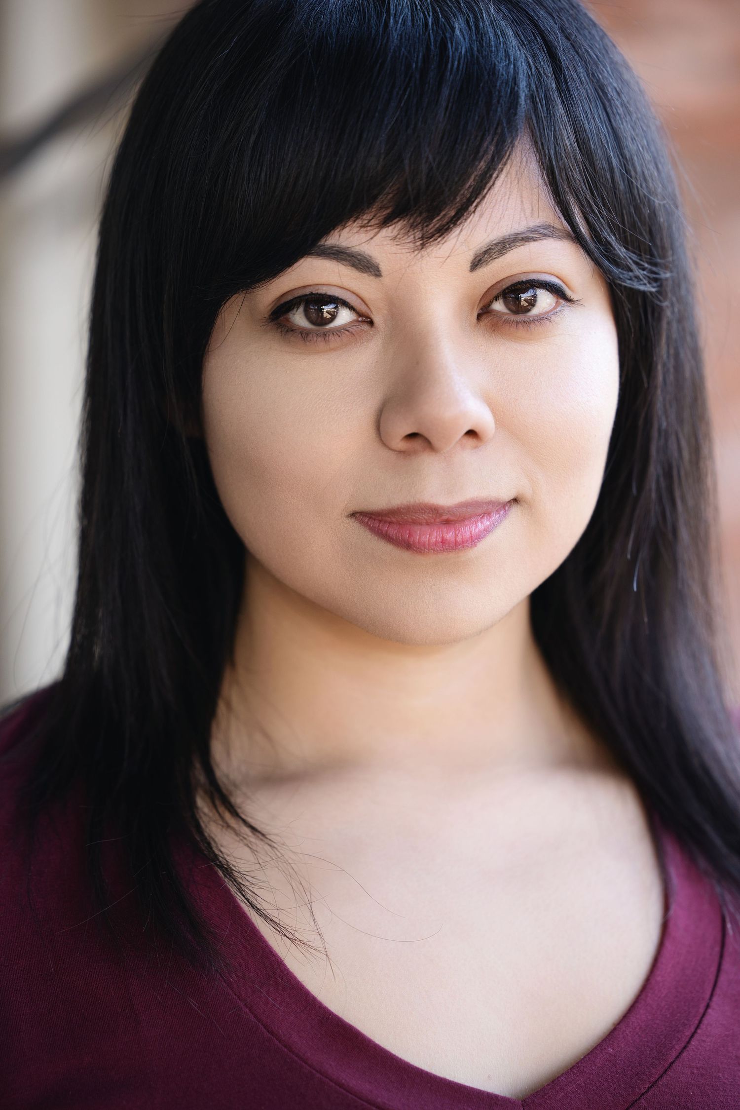 Professional headshot portrait of someone with long black hair wearing a burgundy top against a blurred background.