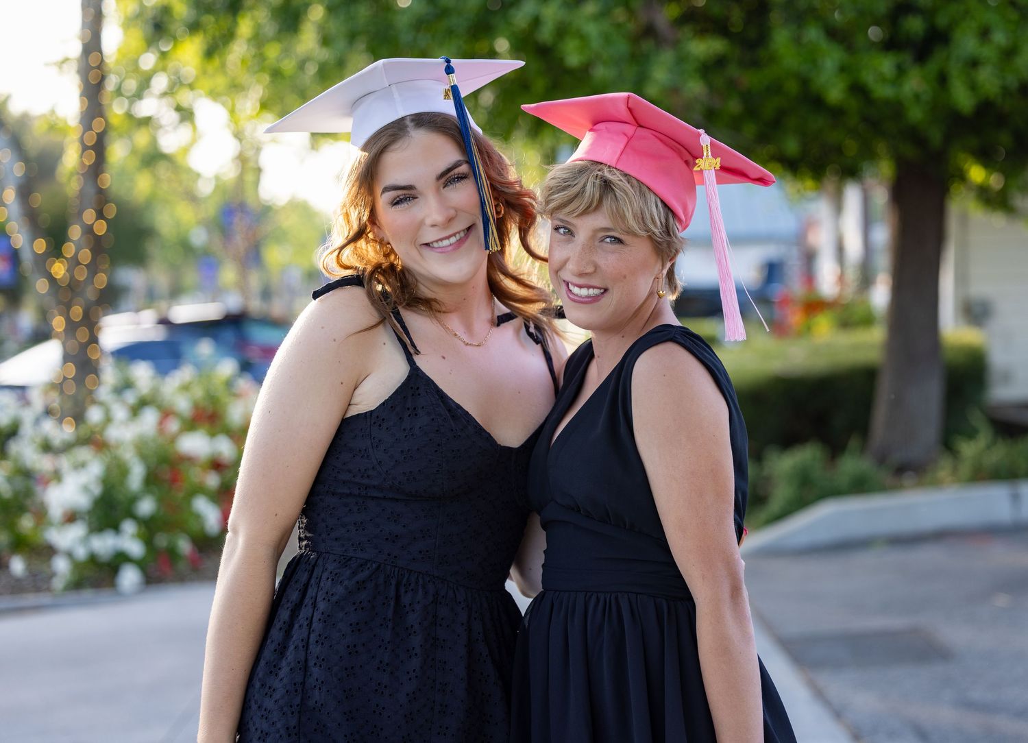 Two people in black dresses and pink graduation caps smile together outdoors on a sunny day.