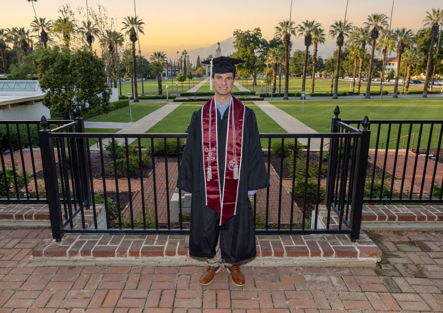 A graduate in cap and gown stands on a brick terrace with palm trees and a sunset sky in the background.