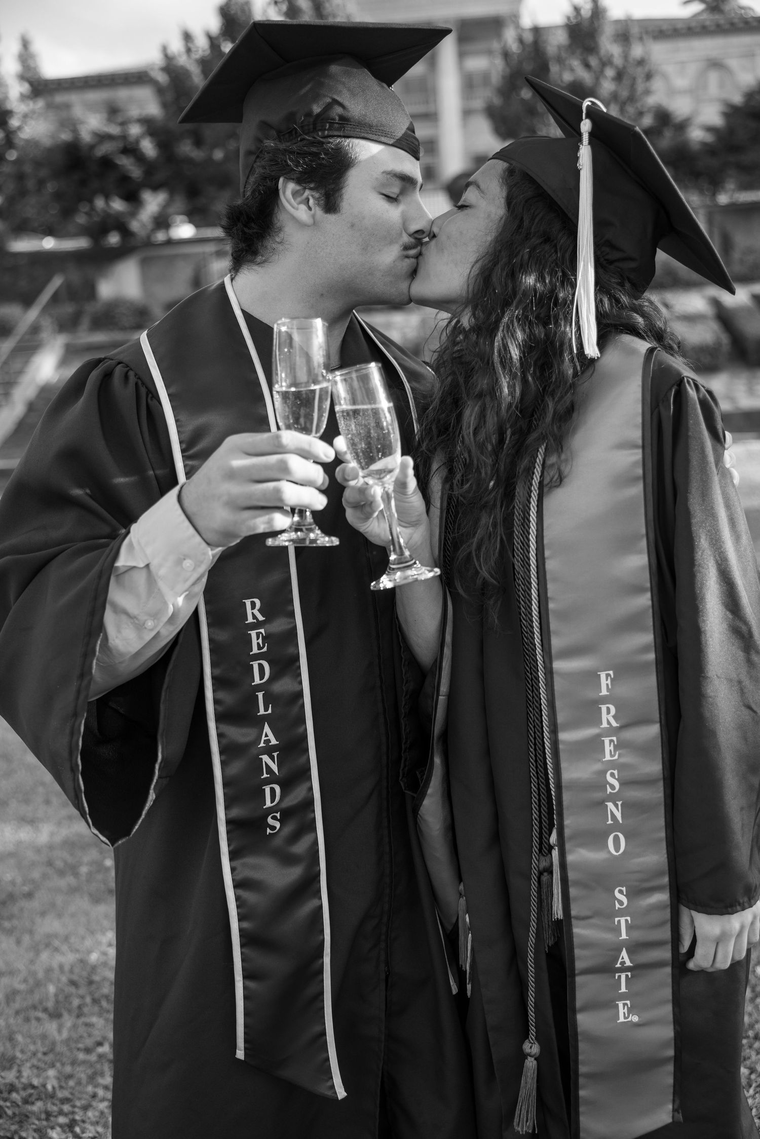 Group of graduates in black gowns and stoles toast with champagne glasses during graduation celebration.