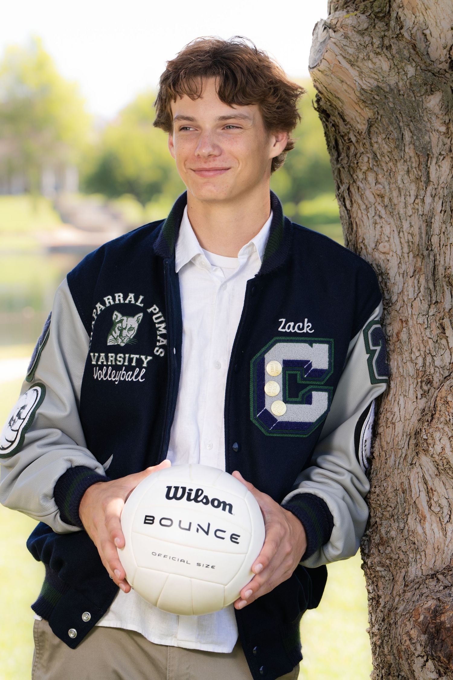 Teen boy in Chaparral Pumas Varsity Volleyball letterman jacket holding Wilson Bounce volleyball outdoors near tree.