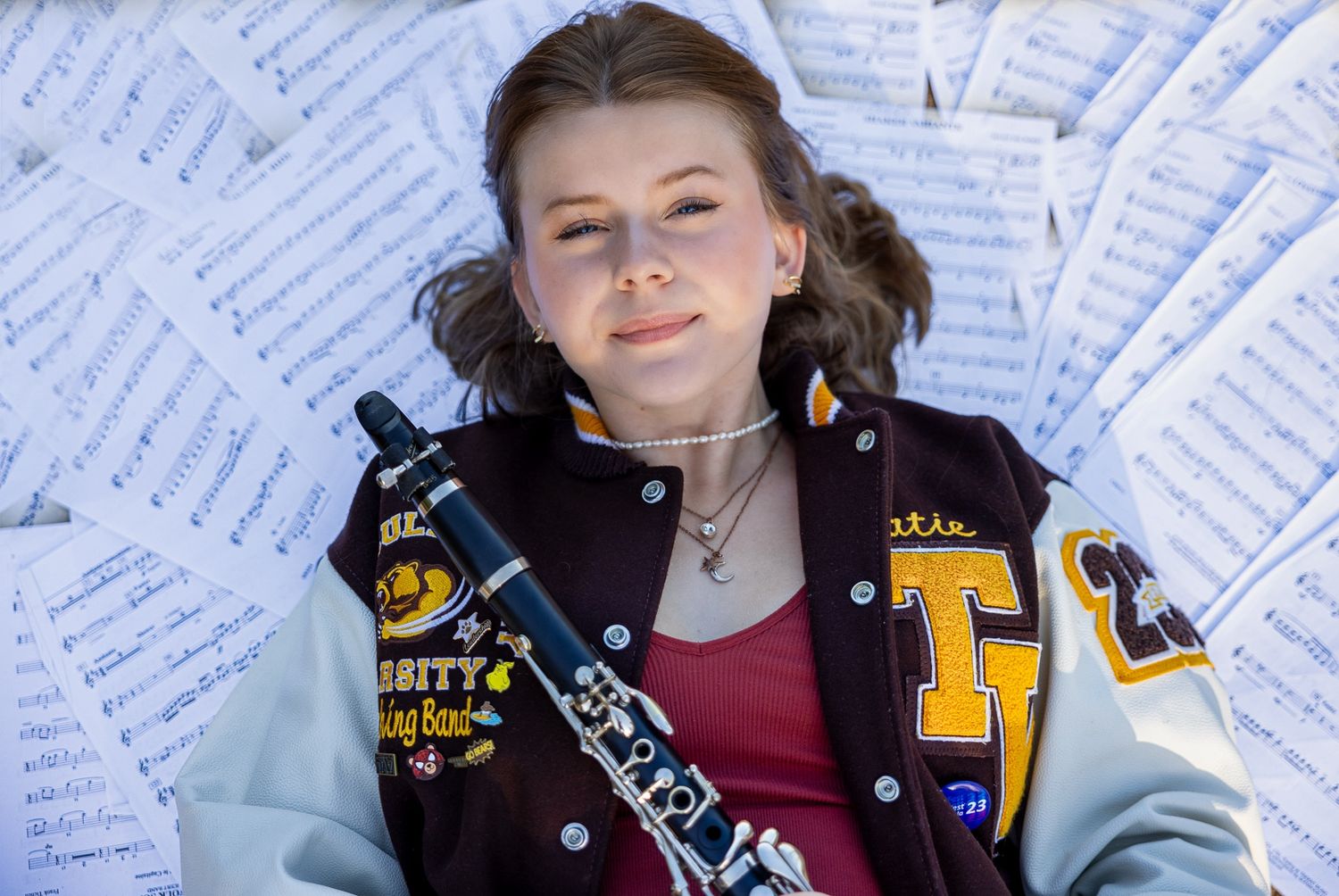 A student wearing a varsity letterman jacket poses with a musical instrument against a background of sheet music.