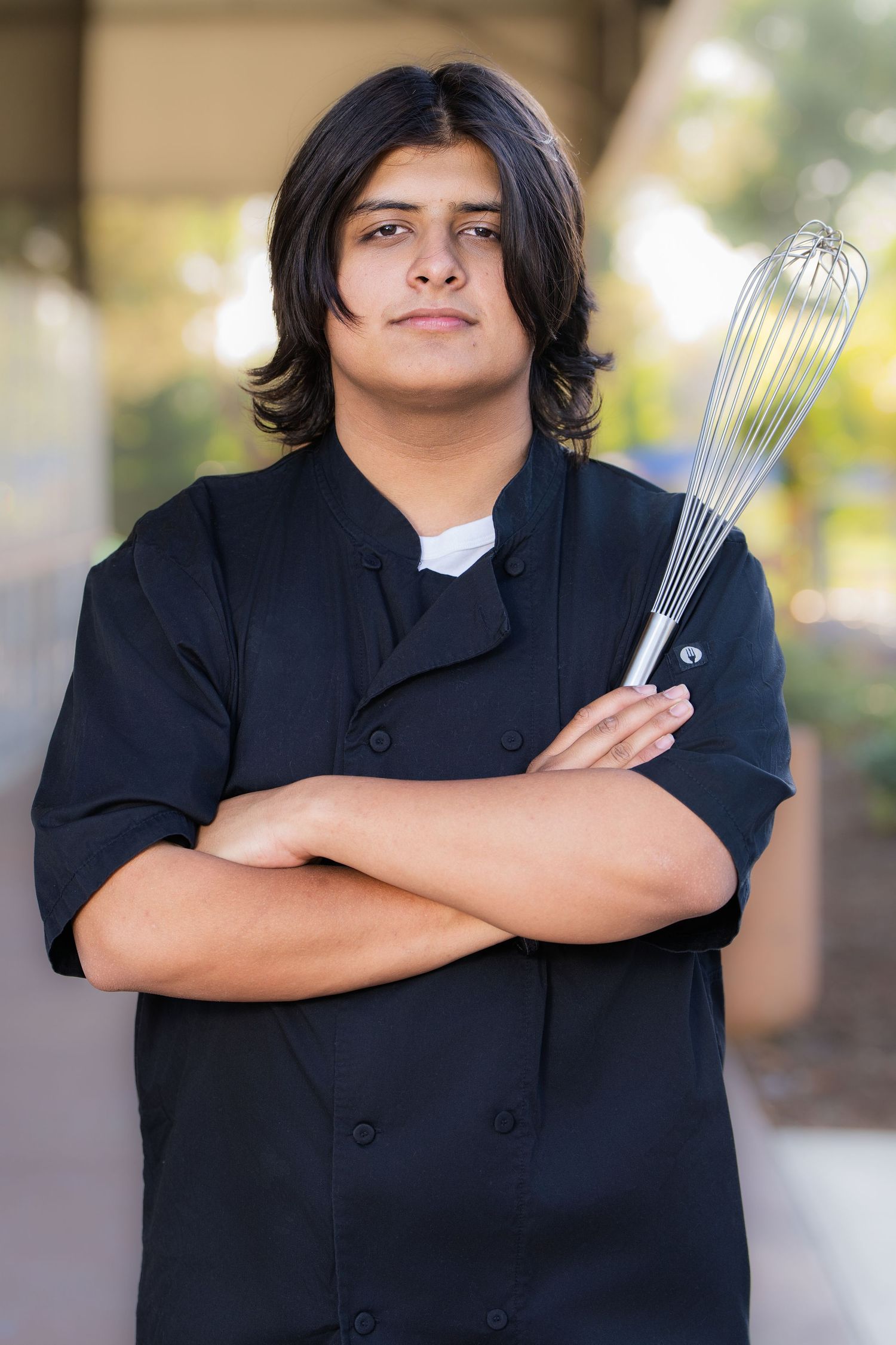 Young chef in black uniform holds a wire whisk with arms crossed, posing confidently outdoors.