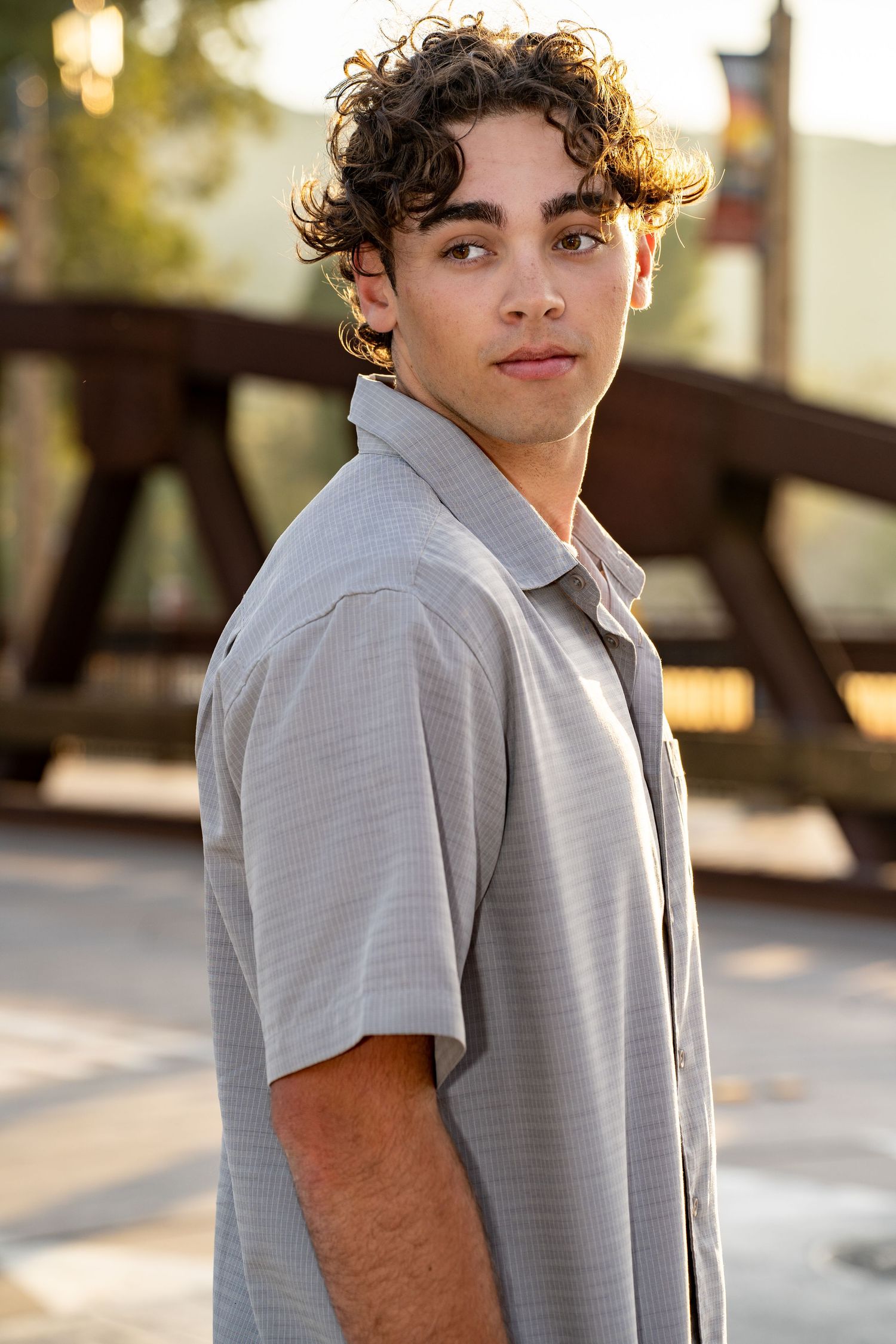 A young person with curly dark hair wearing a light gray button-down shirt poses outdoors at golden hour.