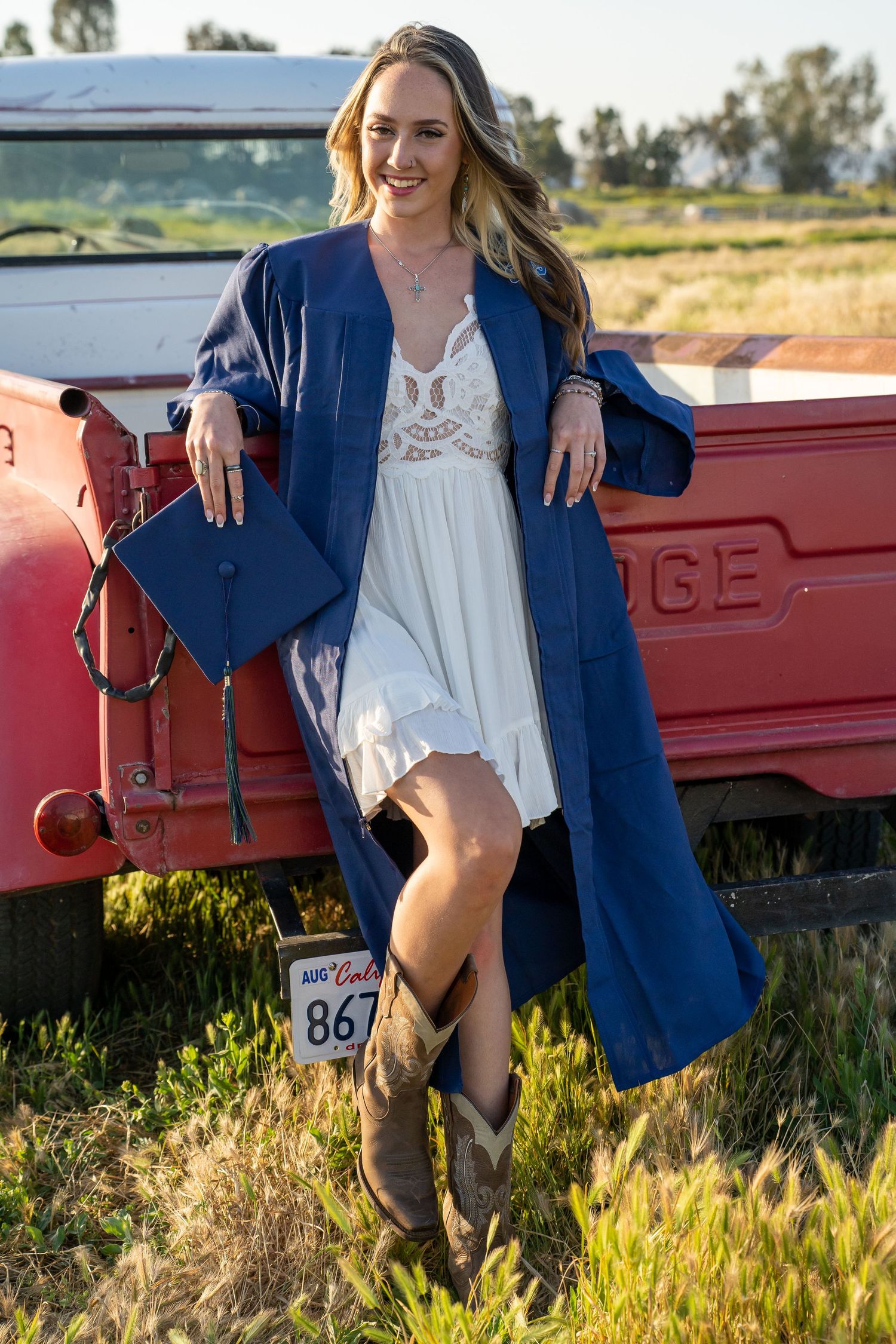 Graduate in blue cap and gown poses with white dress and cowboy boots against vintage red truck in rural setting.