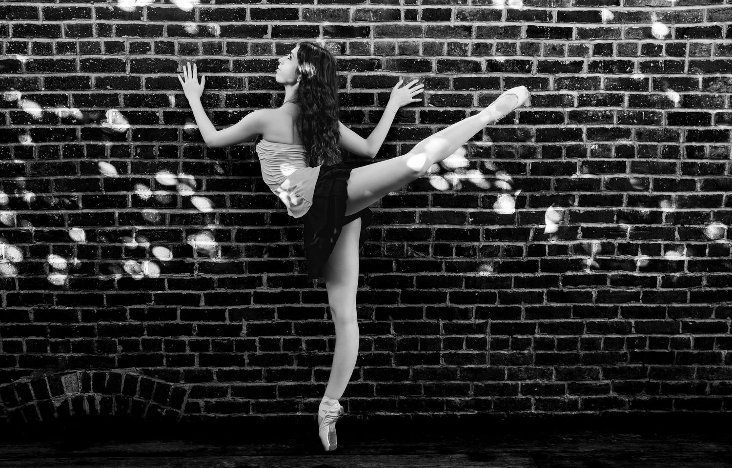 A ballet dancer performs a graceful leap against a dark brick wall in black and white.