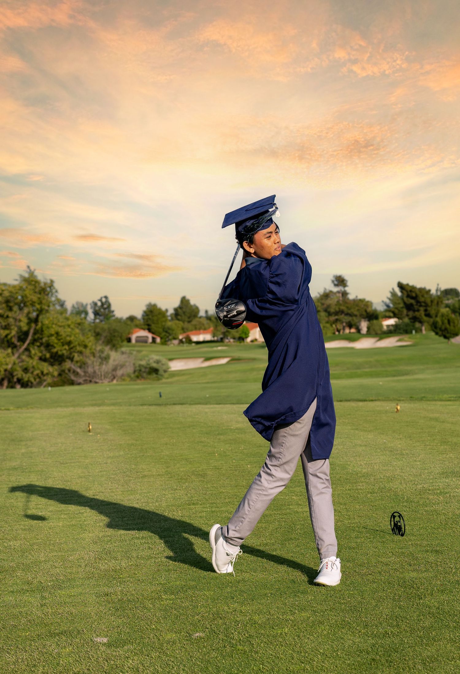 A golfer in graduation attire takes a swing on a golf course during sunset.