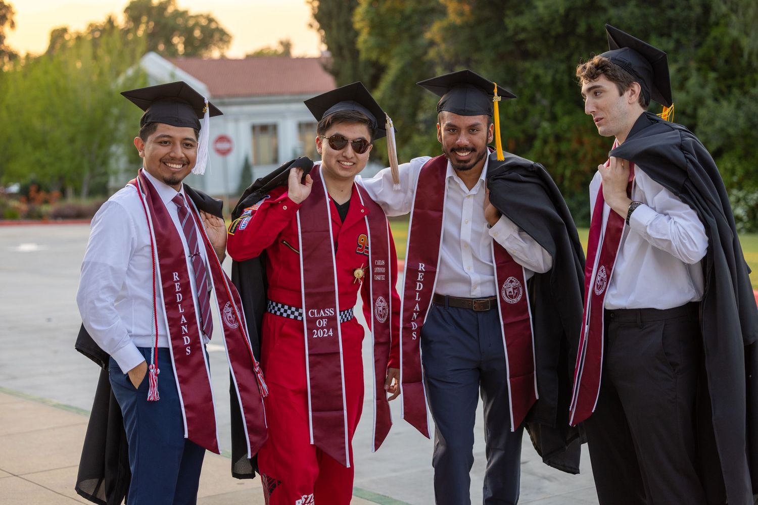 A group of graduates in red and black academic regalia walking and smiling together at sunset.