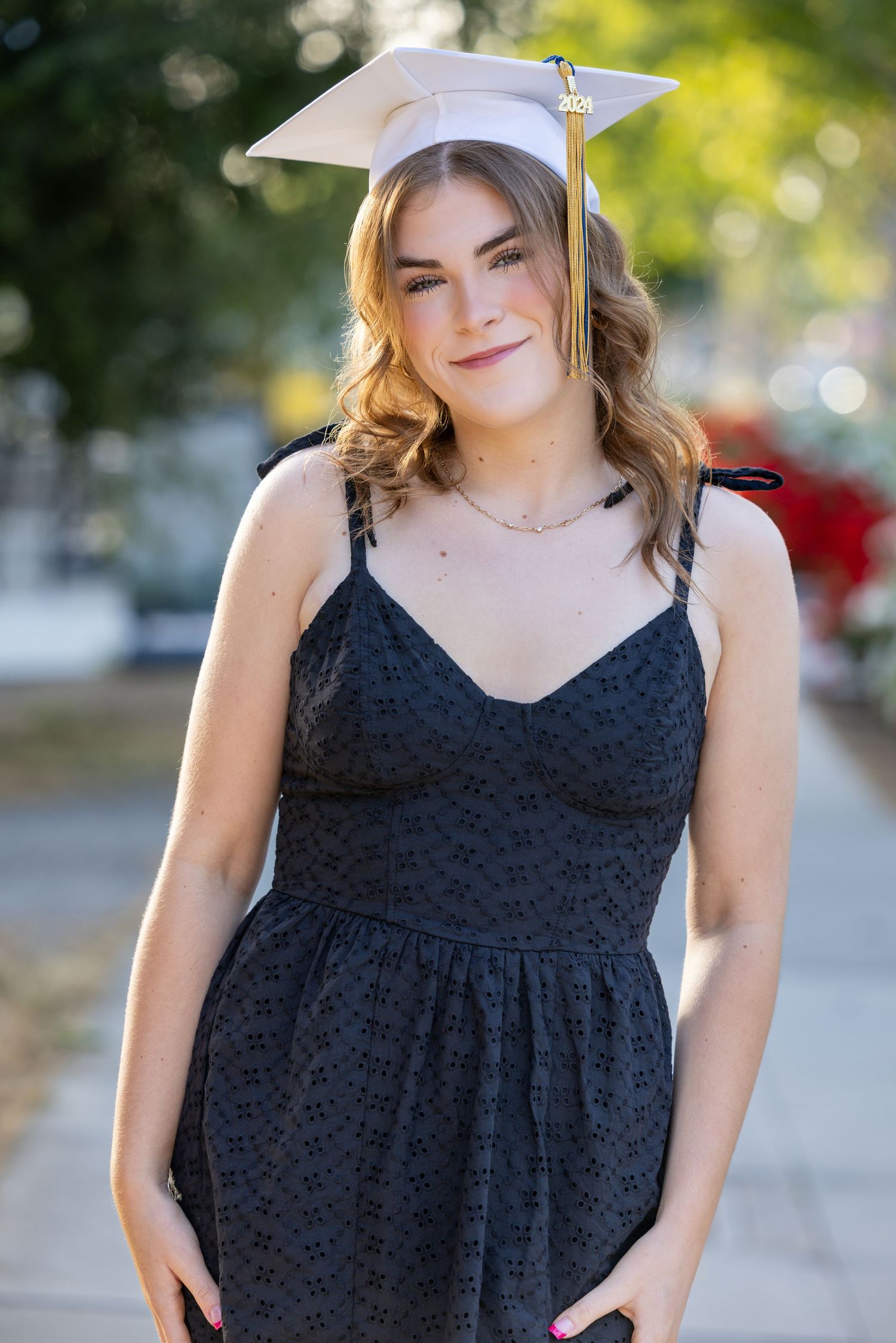 A graduate wearing a black lace dress and cap smiles in an outdoor setting with bokeh background.