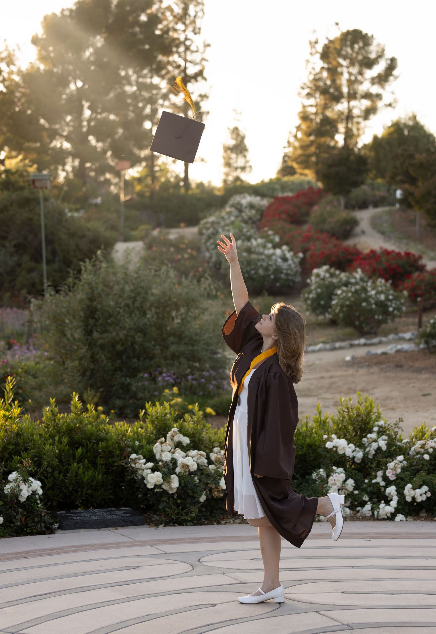 A graduate in cap and gown celebrates on a concrete patio surrounded by lush garden landscaping at sunset.