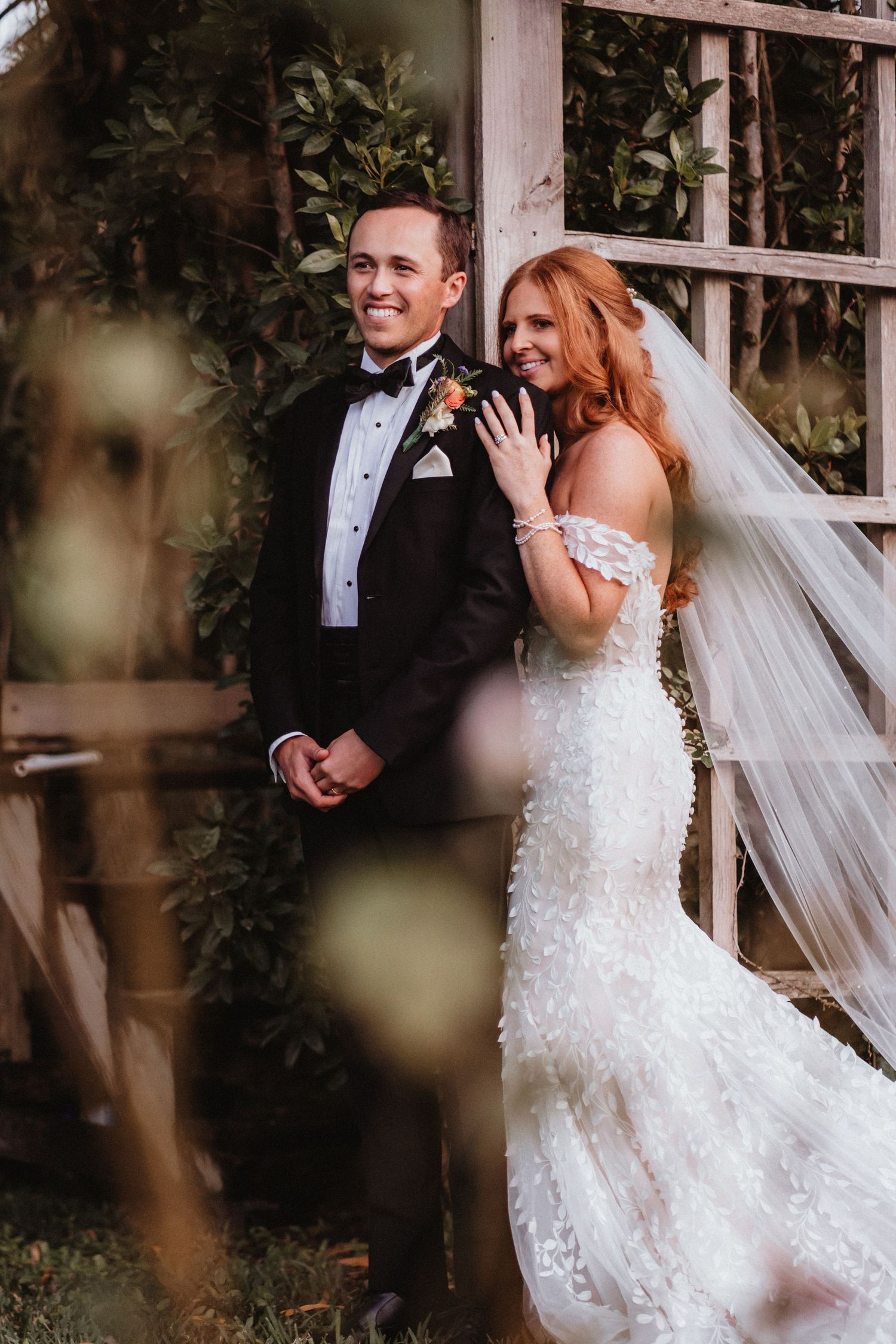 Bride in floral gown and groom in tuxedo pose by a garden trellis.